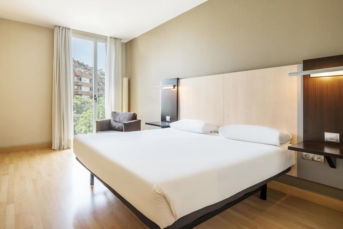 A hotel room with a large bed, a desk, a chair, a nightstand, and a view of buildings through the window.
