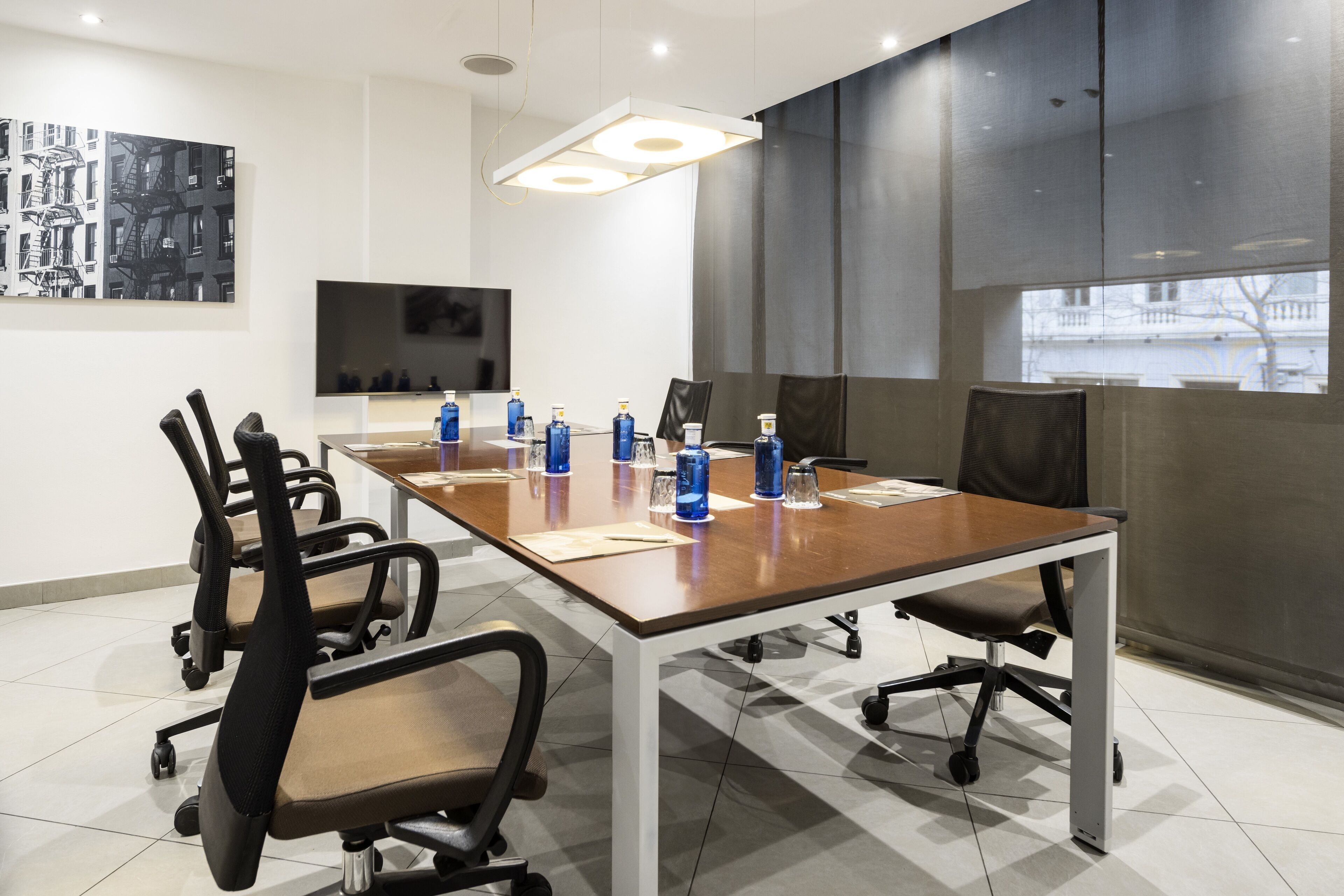 A conference room with a long table, multiple chairs, a flat-screen TV, and a large window with a view of buildings.