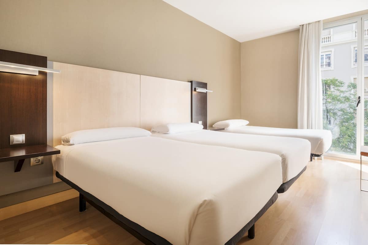 A hotel room with two beds, a desk, and a window with a view of buildings.
