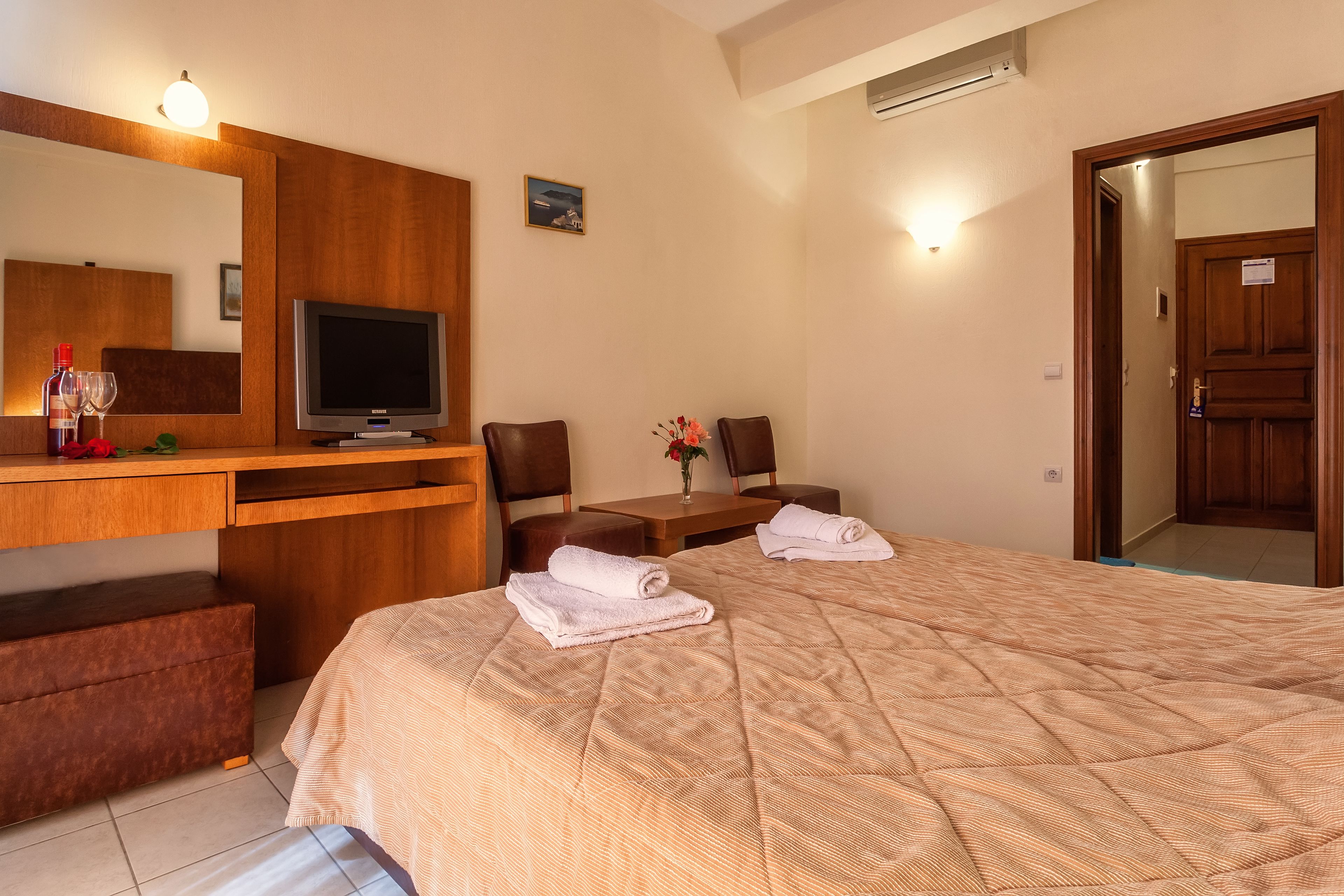 A hotel room with a bed, a desk with a television, a chair, and a small table with a vase of flowers.