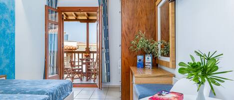 A room with a bed, a desk with books, a plant, and a view through an open door to an outdoor area.