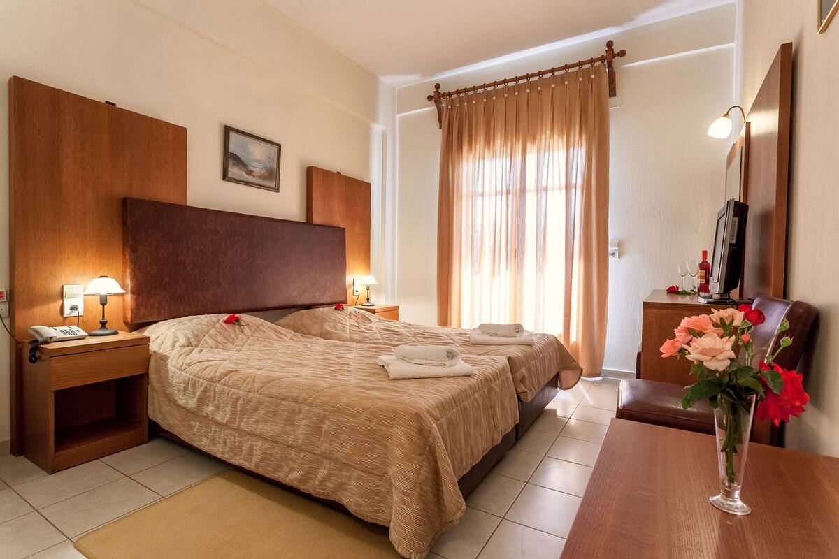 A hotel room with a large bed, bedside lamps, a wooden headboard, a small table with a vase of flowers, and a television on a wooden cabinet.