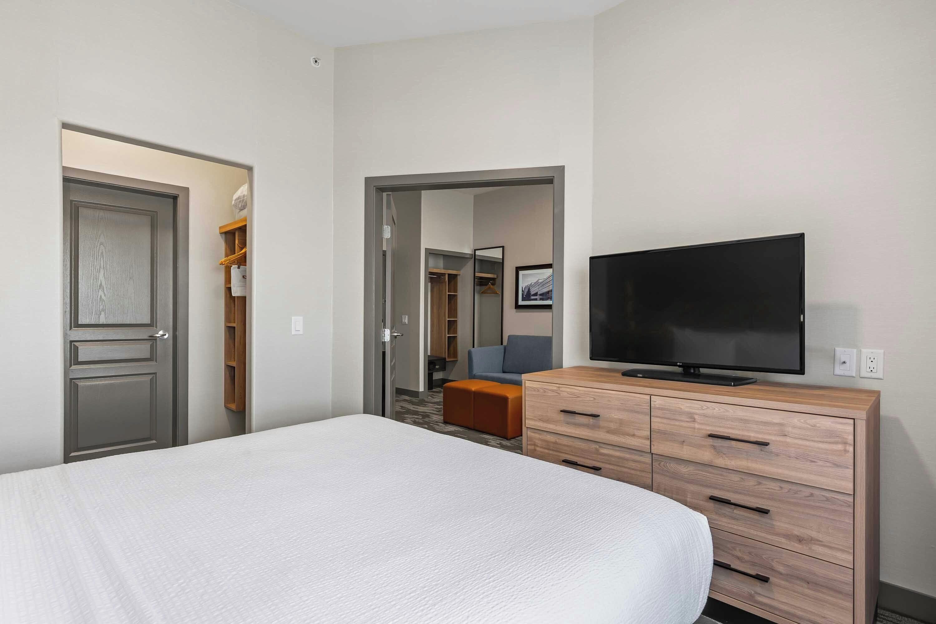 A hotel room with a bed, a television, a wooden dresser, and a closet with hanging clothes.