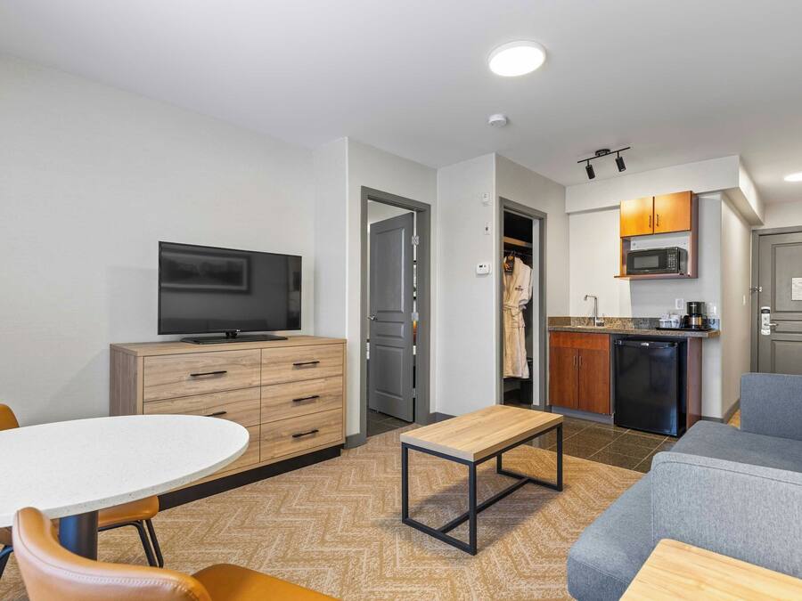 A modern living room with a TV, wooden furniture, a grey sofa, and a kitchen area with a microwave and refrigerator.