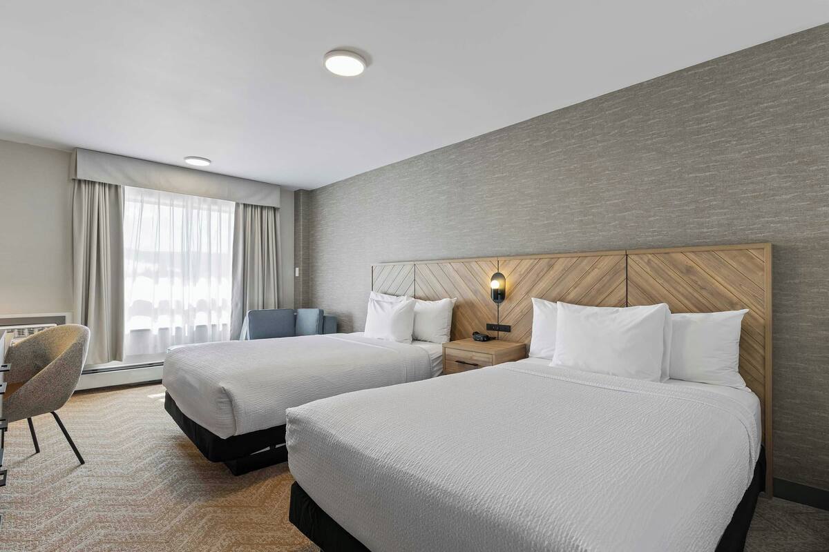 A hotel room with two beds, a desk, a chair, a window with curtains, and a wooden headboard.