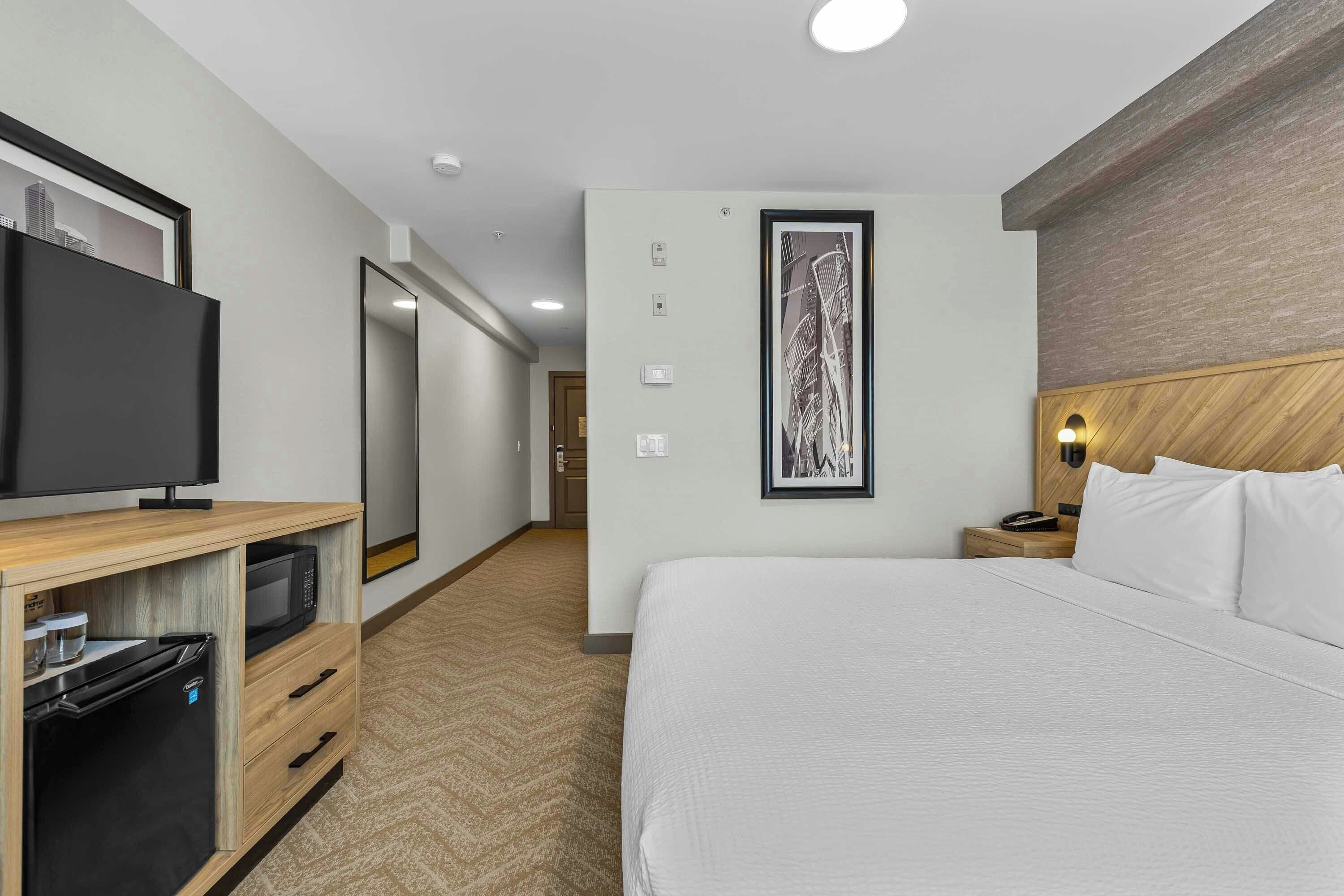 Habitación de hotel con cama, televisión, microondas, refrigerador, mesita de noche y un cuadro en la pared.