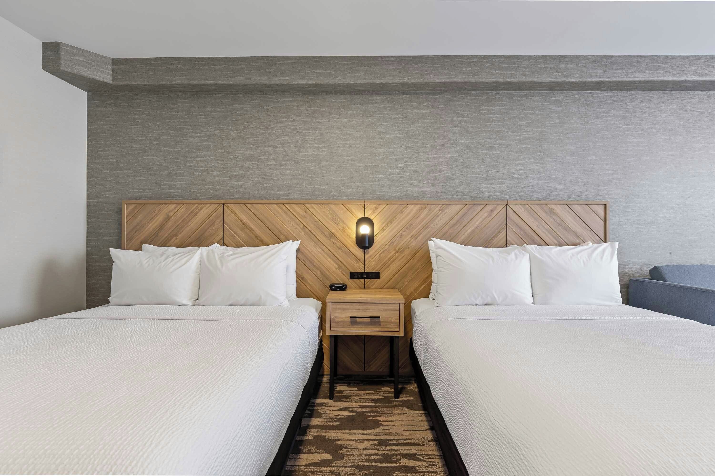 A hotel room with two beds, a wooden headboard, a nightstand with a lamp, and a patterned rug.