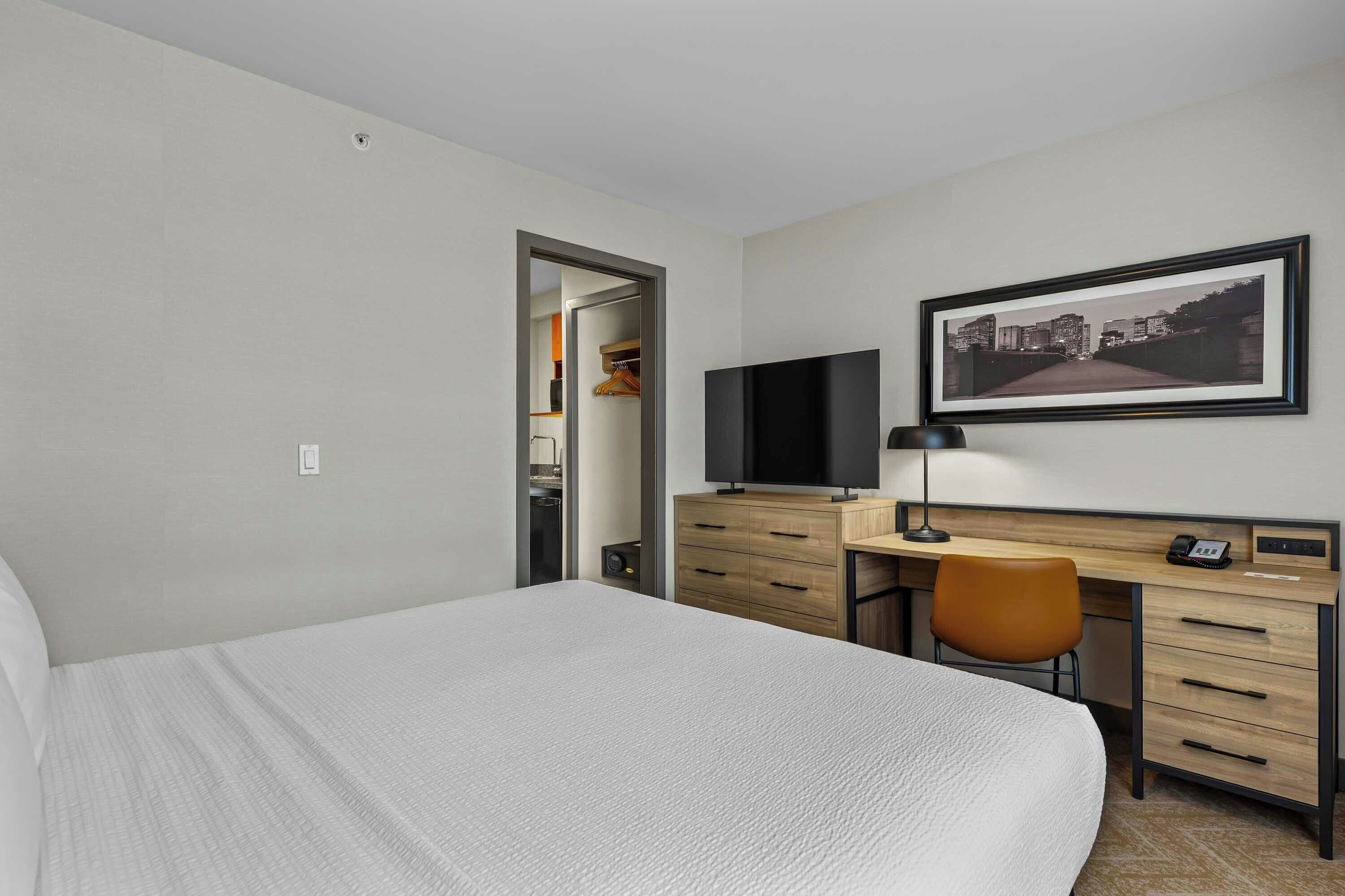 A hotel room with a bed, a desk with a computer, a chair, a television, and a framed picture on the wall.