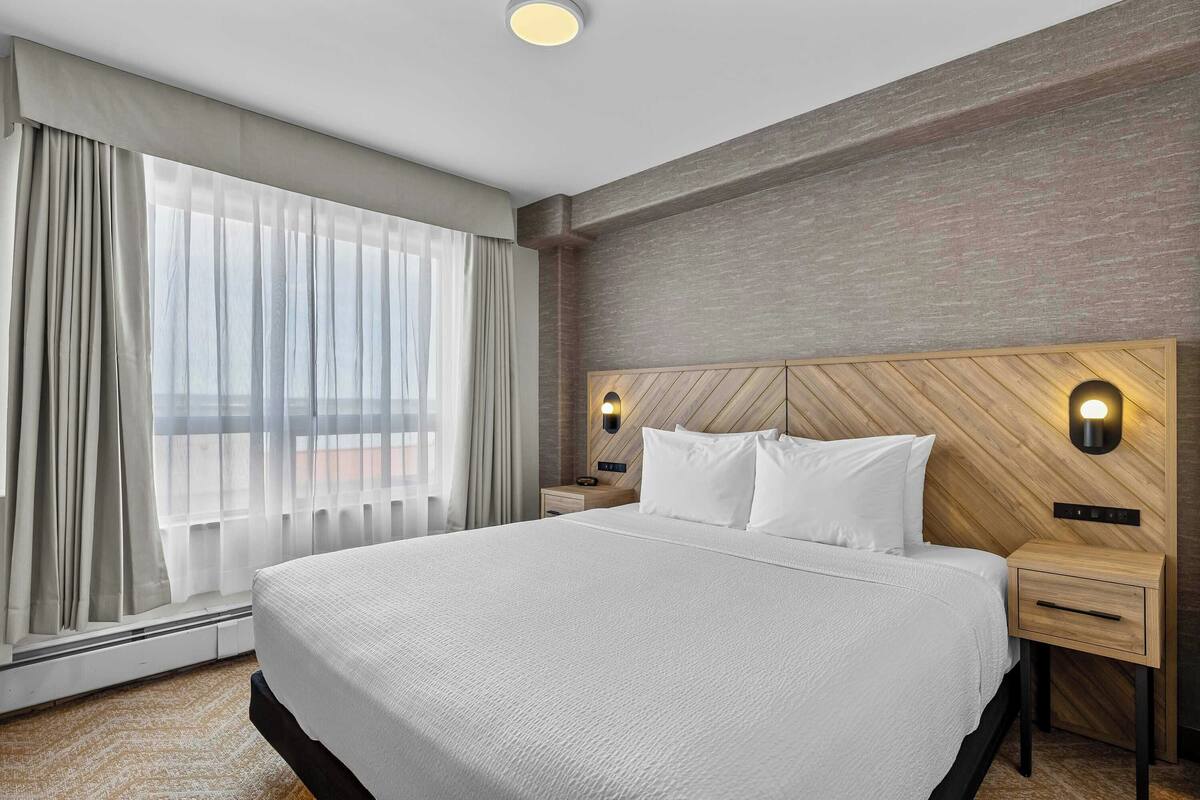 A hotel room with a large bed, two bedside tables, a wooden headboard, a window with sheer curtains, and a wall-mounted light fixture.