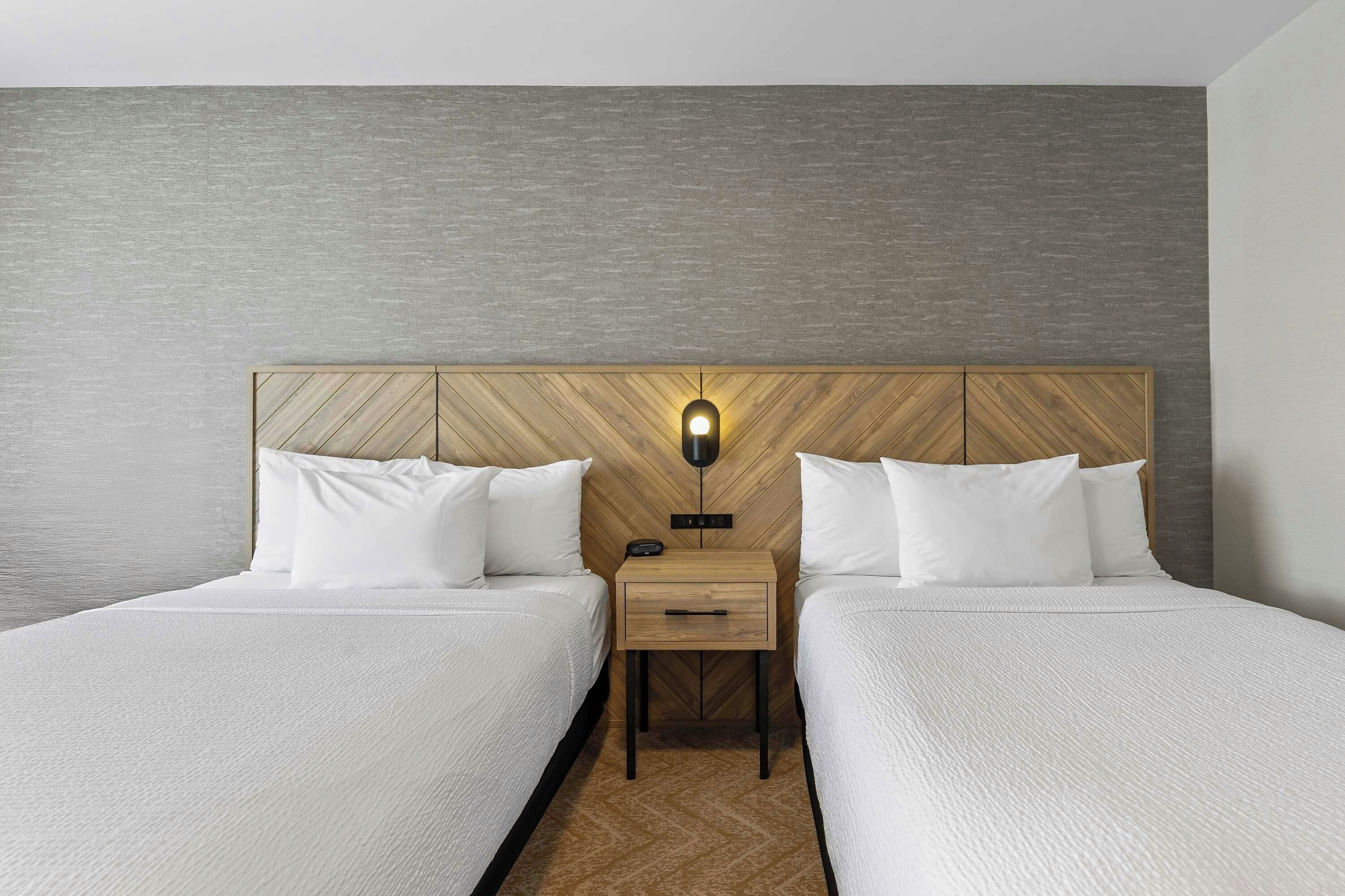A hotel room with two beds, a nightstand with a lamp, and a wooden headboard.