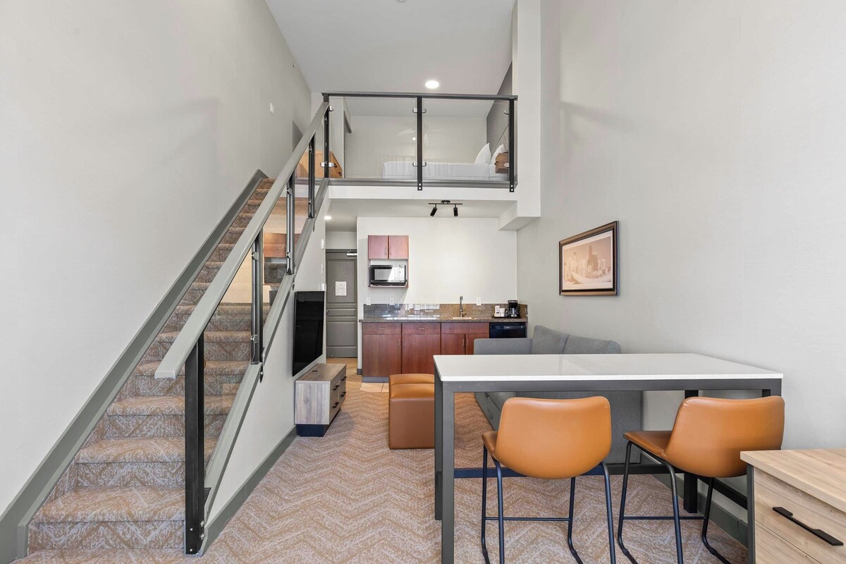 A modern interior with a staircase, a dining area with two chairs, a kitchen with wooden cabinets, and a loft bedroom with a bed.