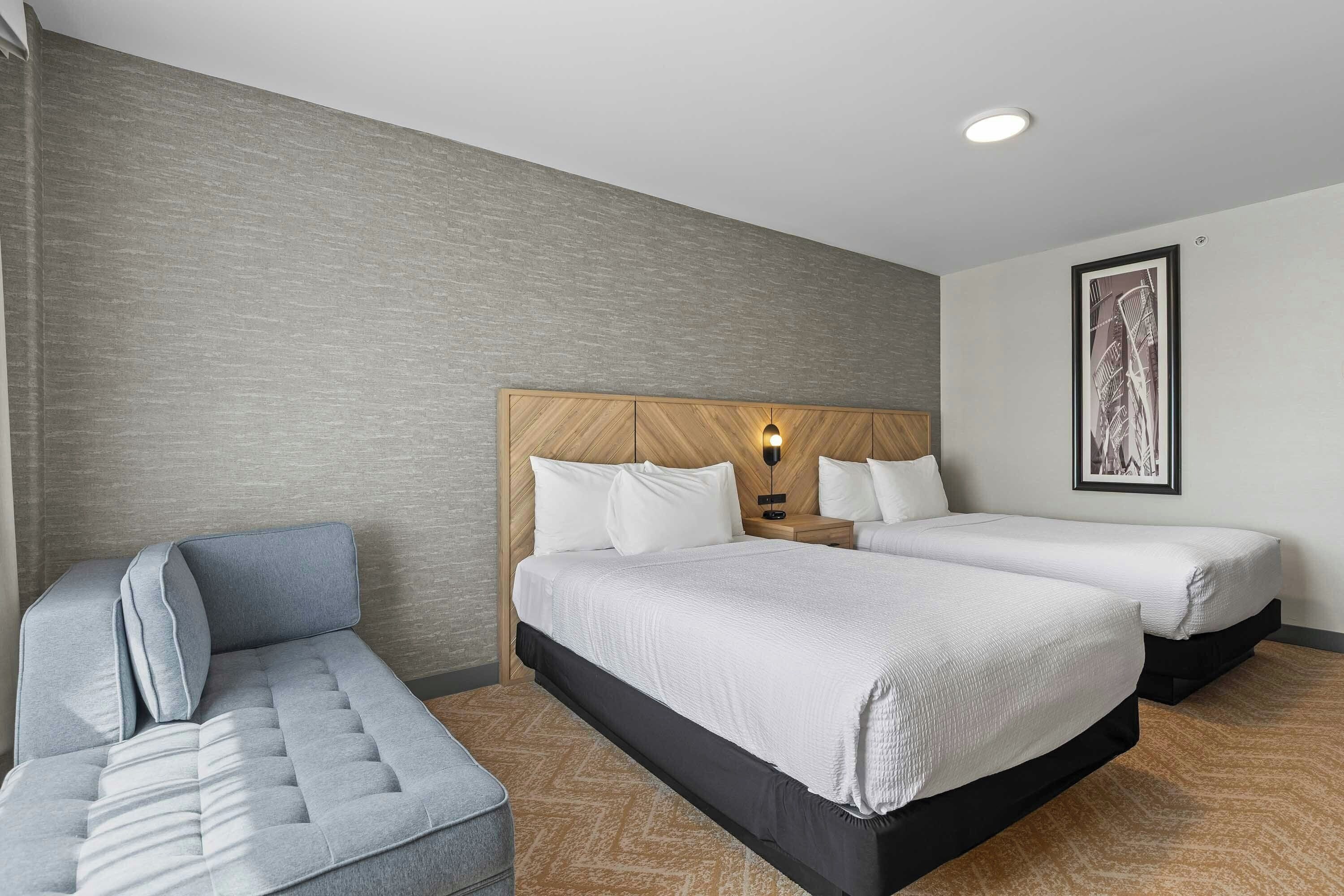 A hotel room with two beds, a sofa, a wooden headboard, a wall-mounted light, and a framed picture.