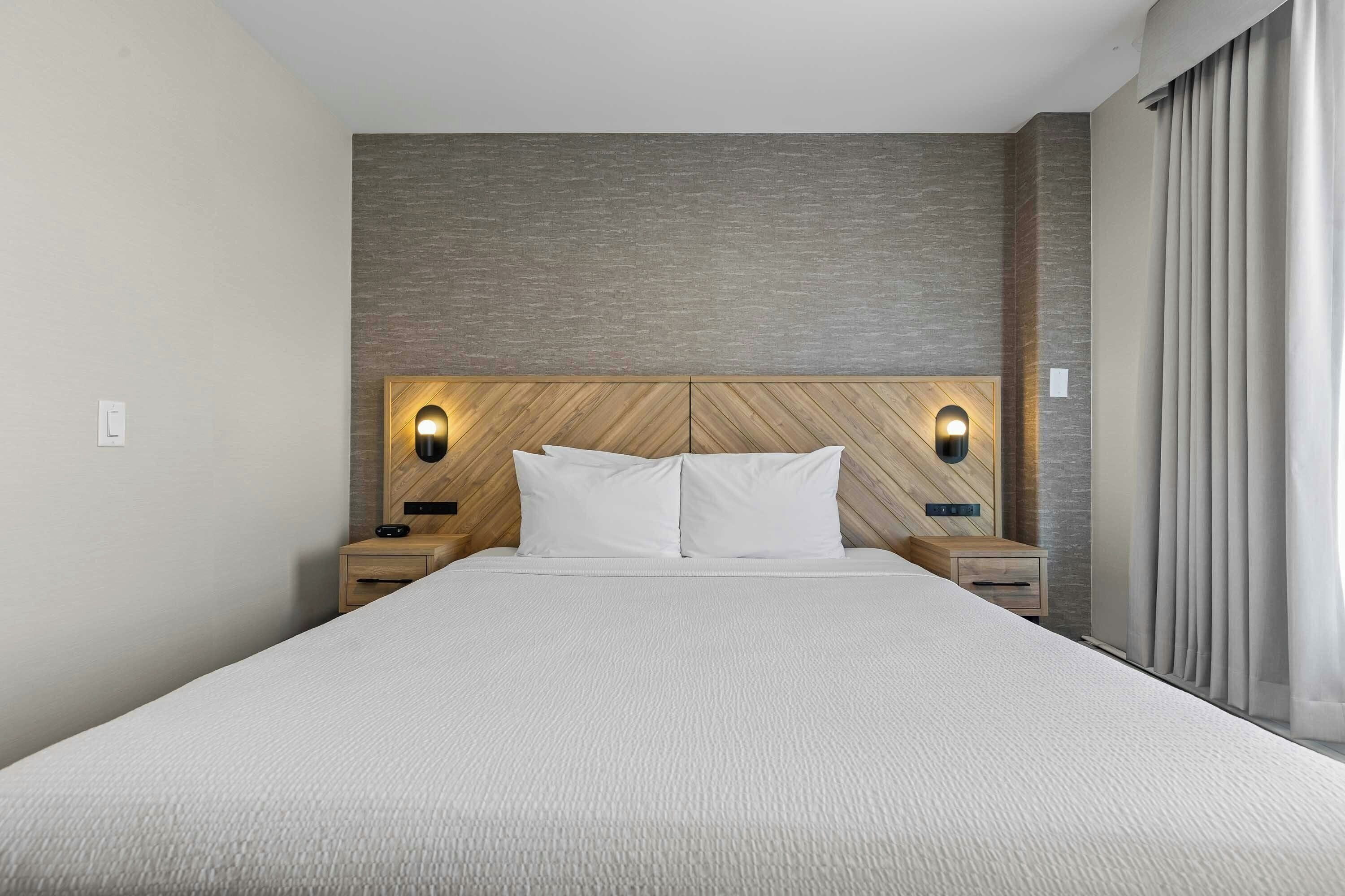 Habitación de hotel con una cama grande, cabecero de madera, dos mesitas de noche y una lámpara fijada a la pared.