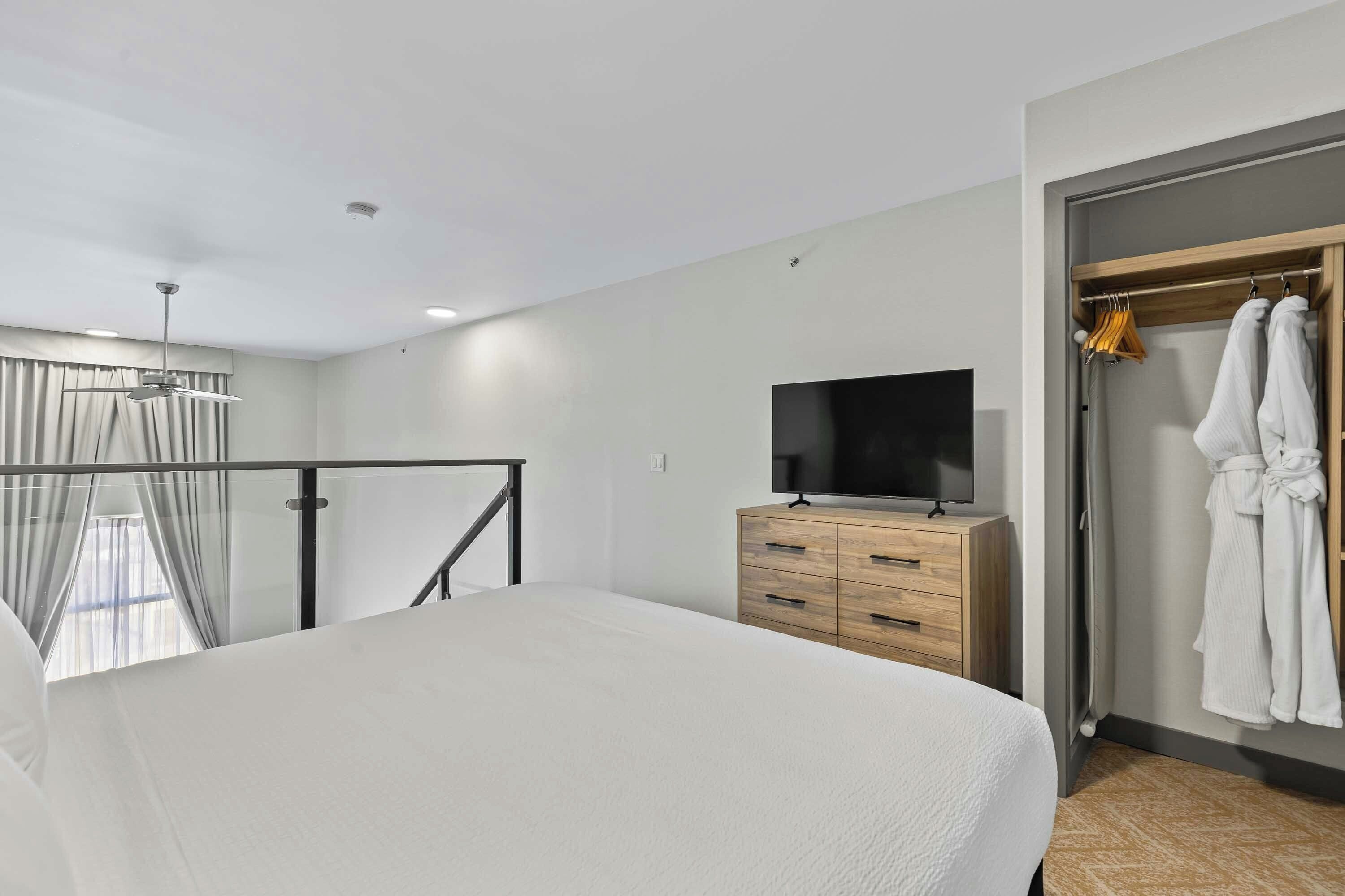 A bedroom with a bed, a TV on a wooden dresser, a wardrobe, and a balcony with curtains.