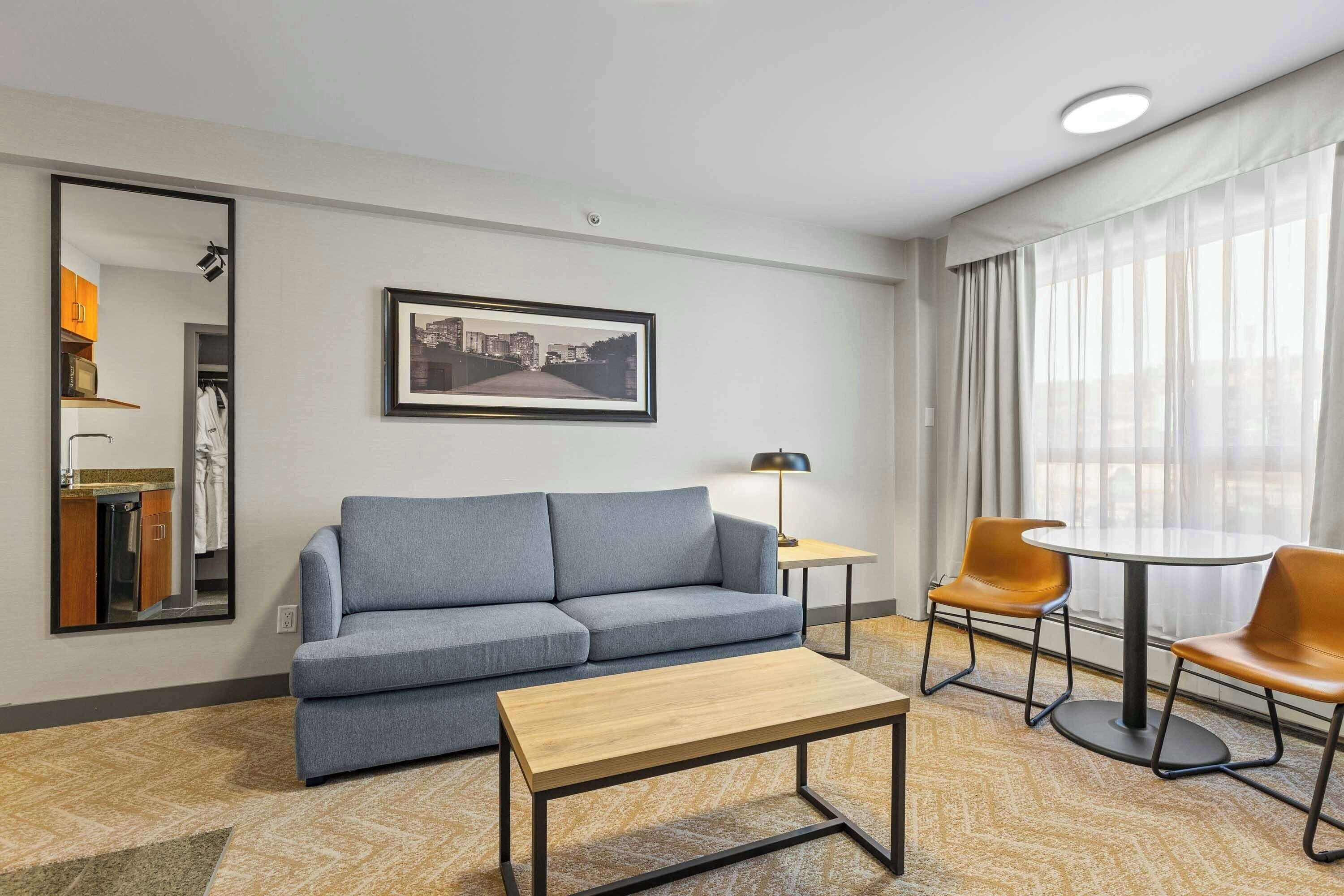 Una habitación de hotel moderna con un sofá gris, una mesa de centro de madera, dos sillas naranja, una mesita con una lámpara y un ventanal grande con cortinas translúcidas.