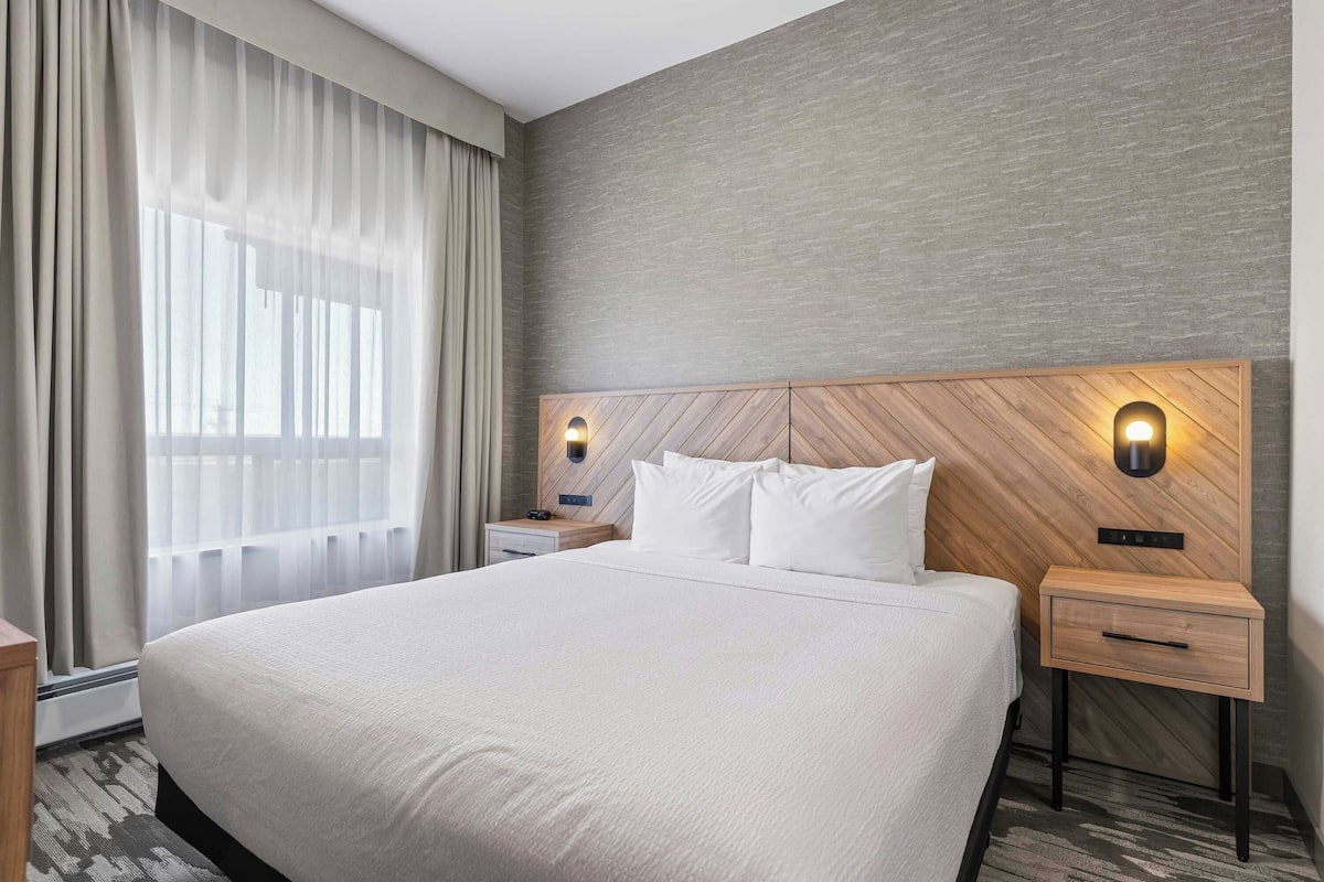 A hotel room with a large bed, two bedside tables, a wooden headboard, and a window with sheer curtains.