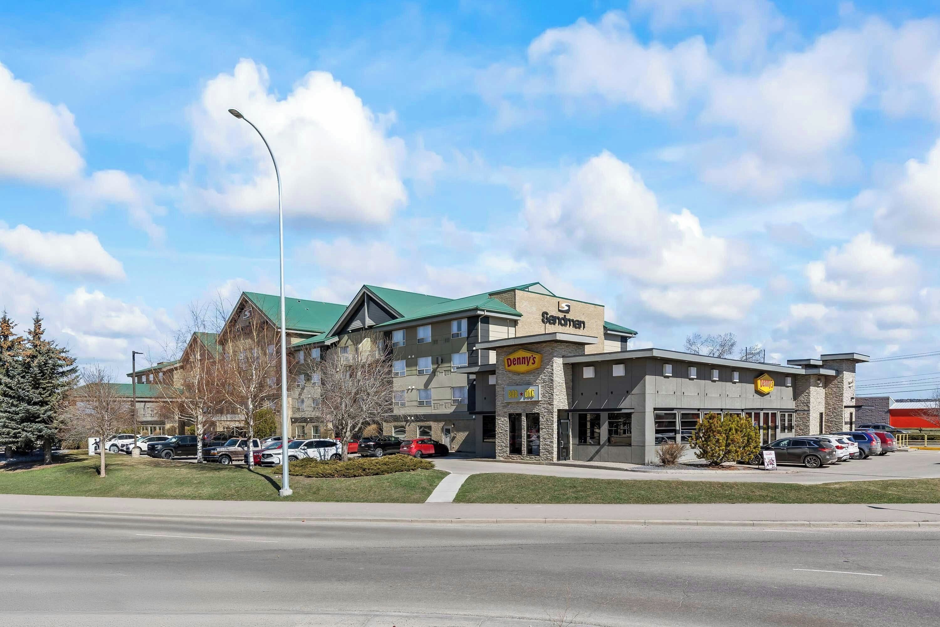 A multi-story building with a green roof, a visible sign for 'Denny's,' and a parking lot with several cars.