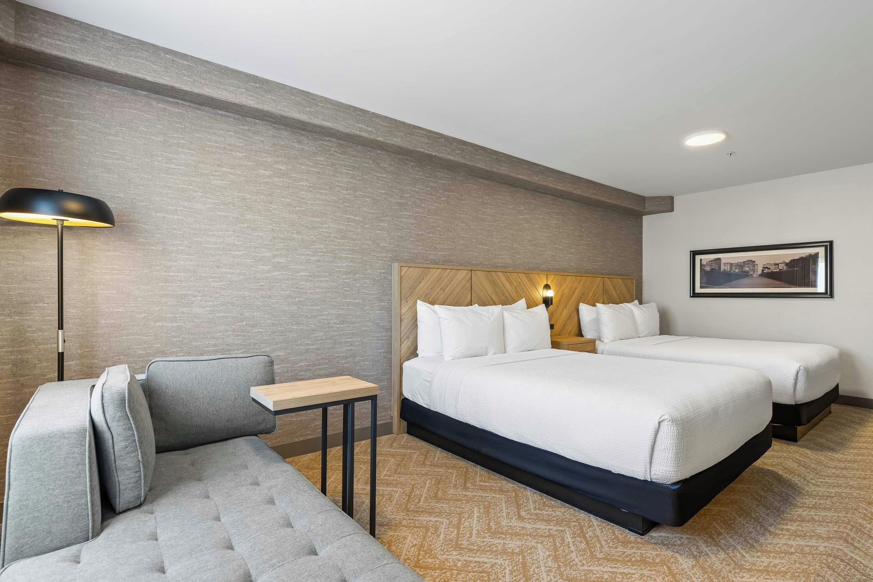 Habitación de hotel con dos camas, un sofá, una mesita, una lámpara y un cuadro en la pared.