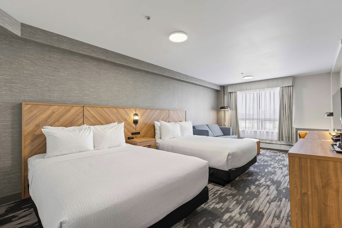 A hotel room with two beds, a desk, a lamp, and a large window with curtains.