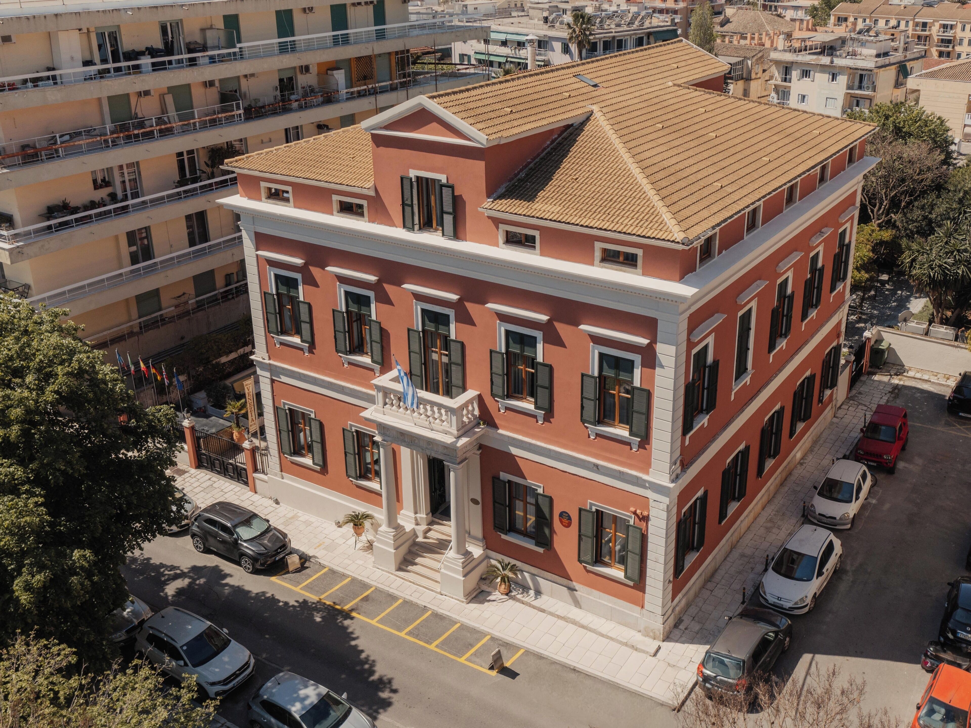A multi-story building with a red facade, balconies, and a parking area with cars.