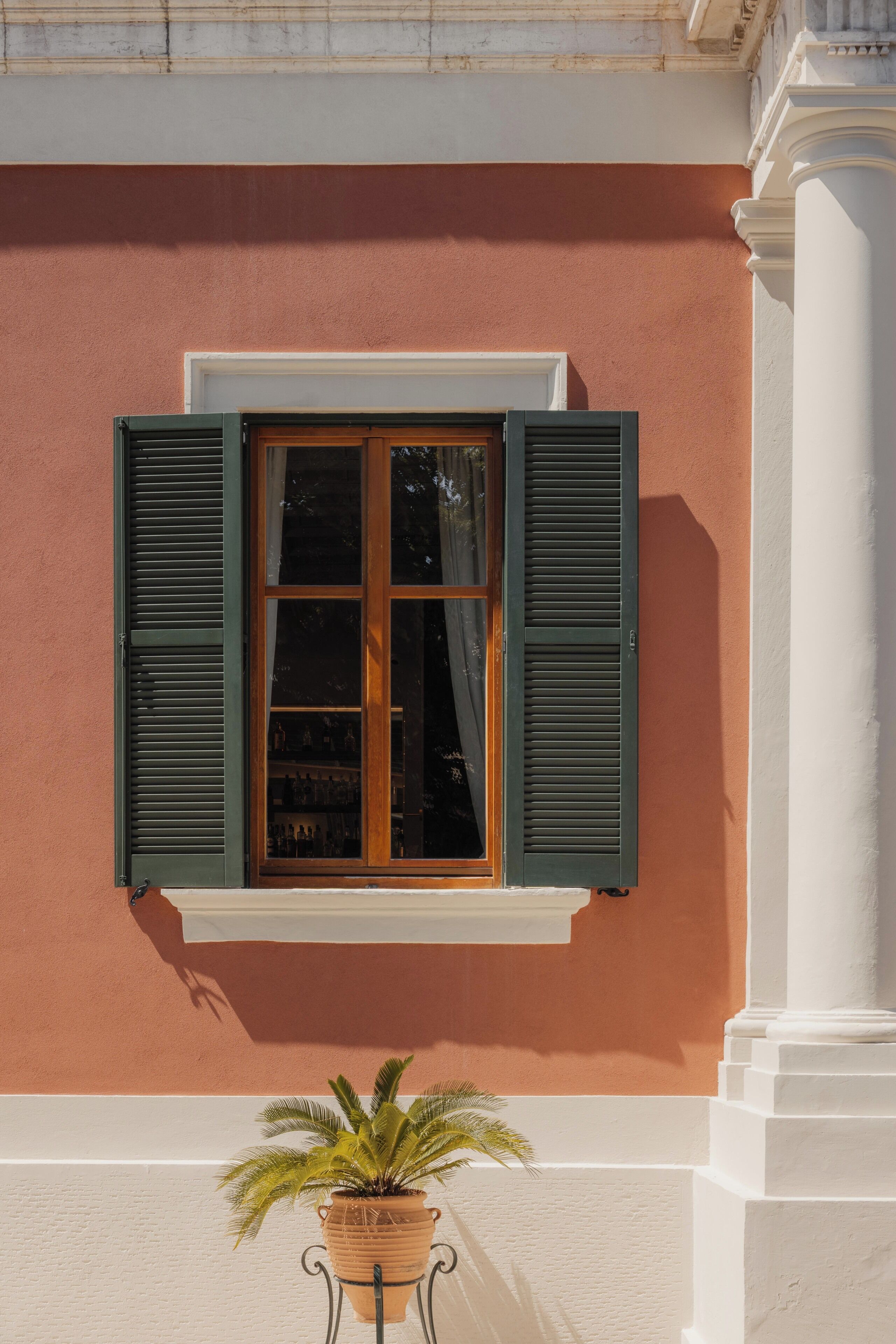 A window with shutters, a potted plant, and a white column.