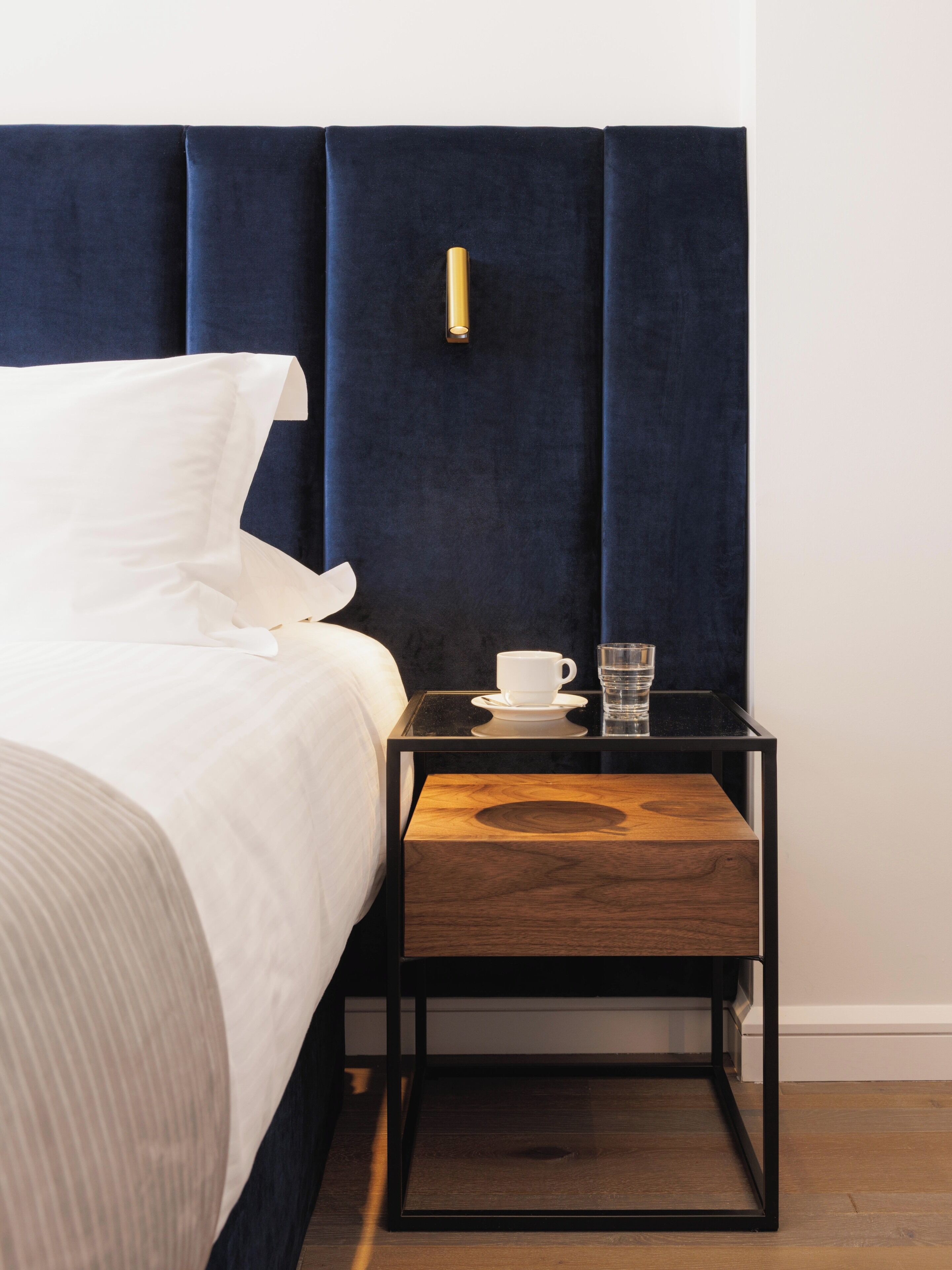A neatly made bed with white linens, a bedside table with a cup and glass, and a dark blue headboard.