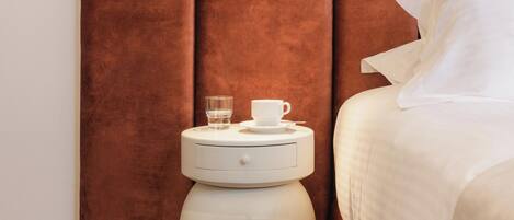 Una cama bien hecha con ropa blanca, un cabecero marrón, una mesita de noche pequeña con una taza y un vaso, y un buró blanco.