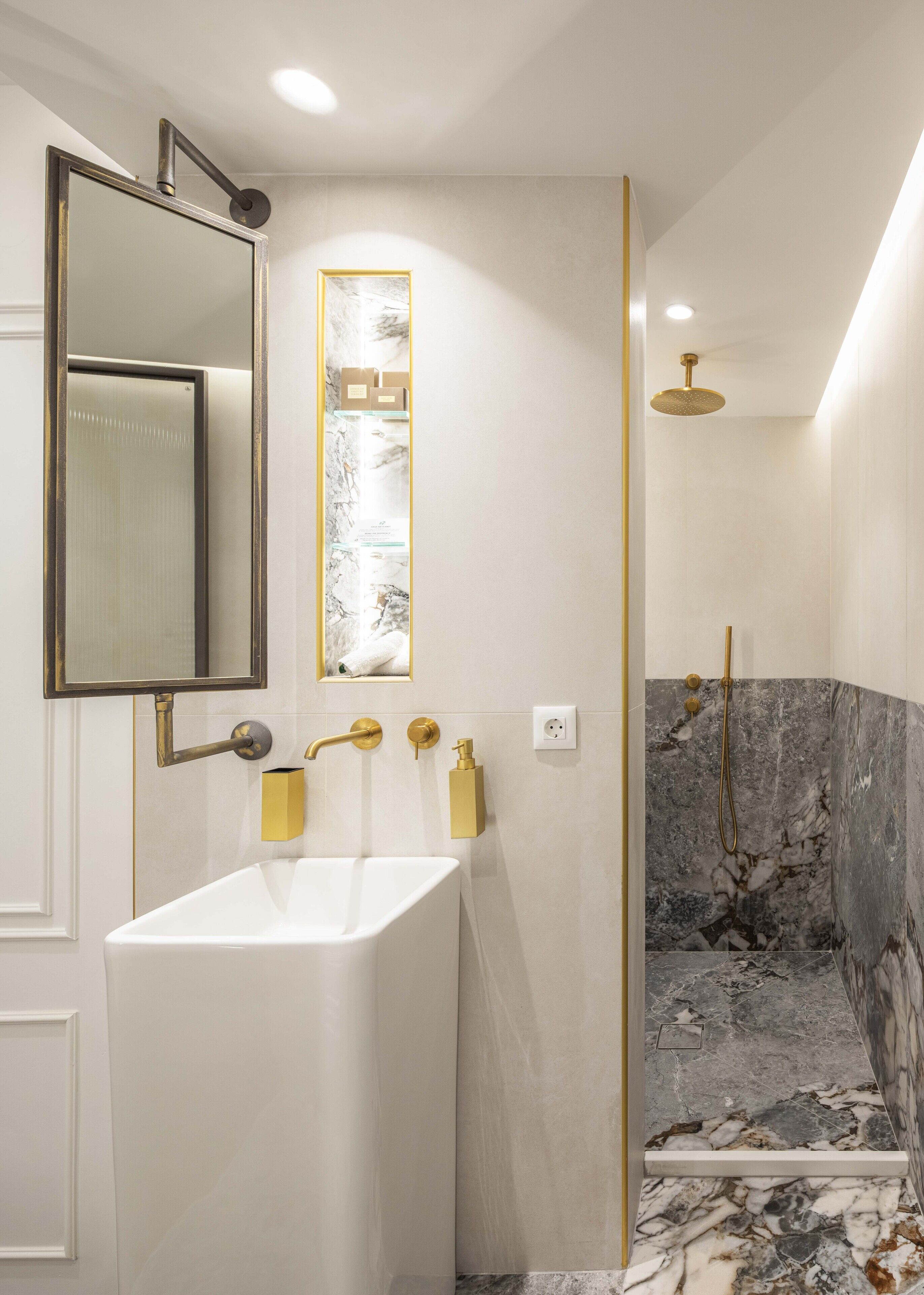 A modern bathroom with a white sink, gold fixtures, a large mirror, and a shower with marble tiles.