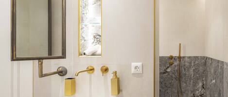 A modern bathroom with a white sink, gold fixtures, a large mirror, and a shower with marble tiles.
