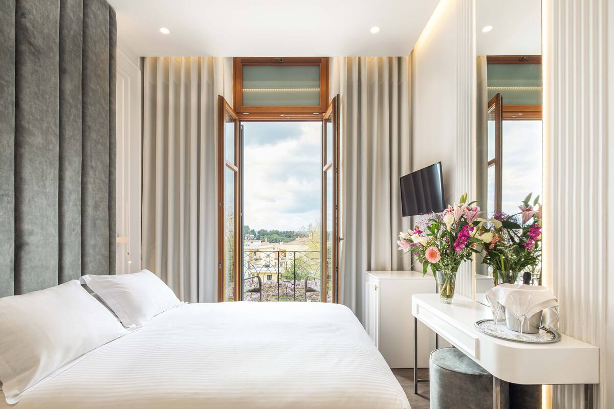 Una habitación de hotel moderna con una cama grande, televisión, escritorio con un jarrón de flores y un balcón con vistas.