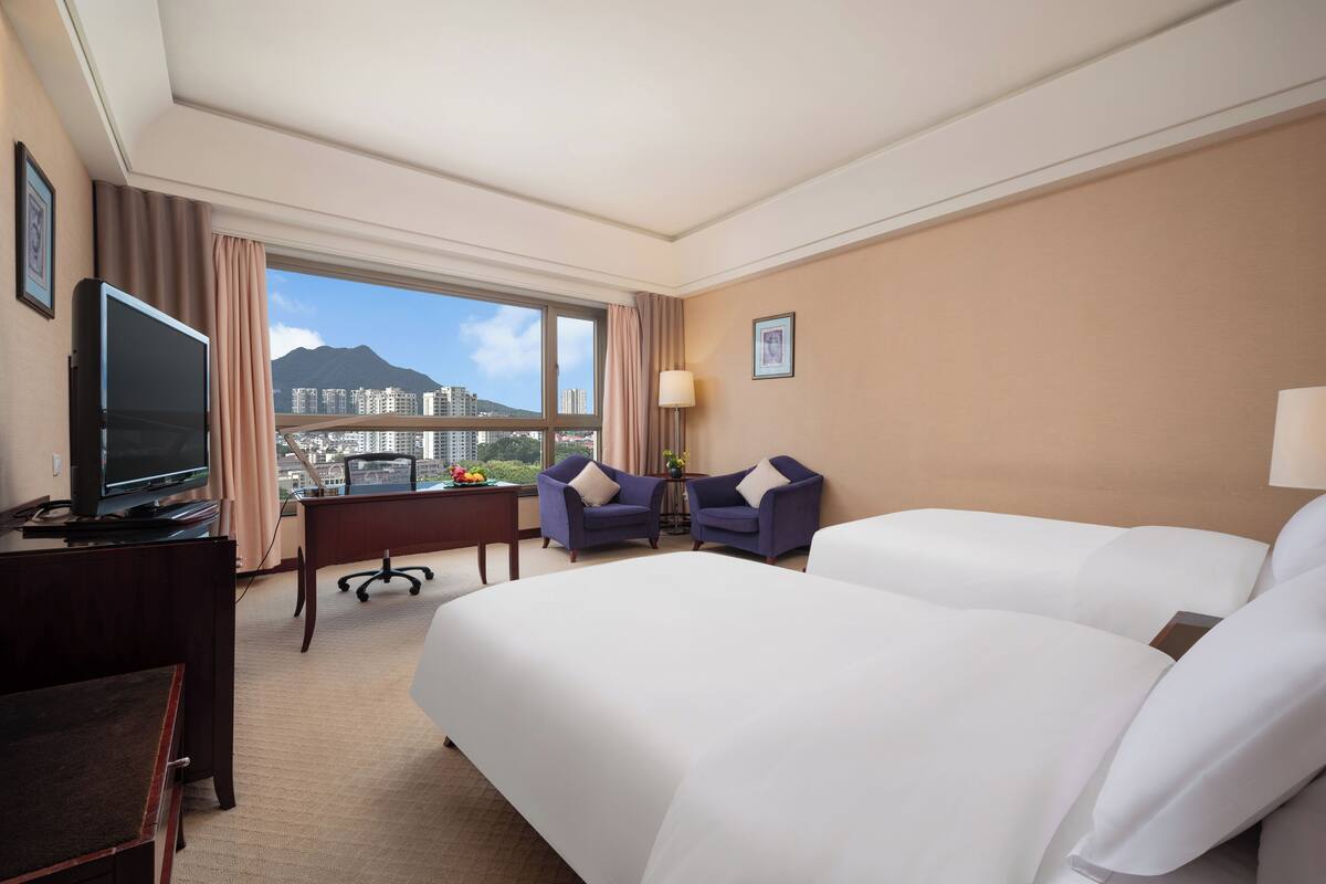 Habitación de hotel con dos camas, un escritorio con televisor, una silla y vistas a un paisaje urbano.