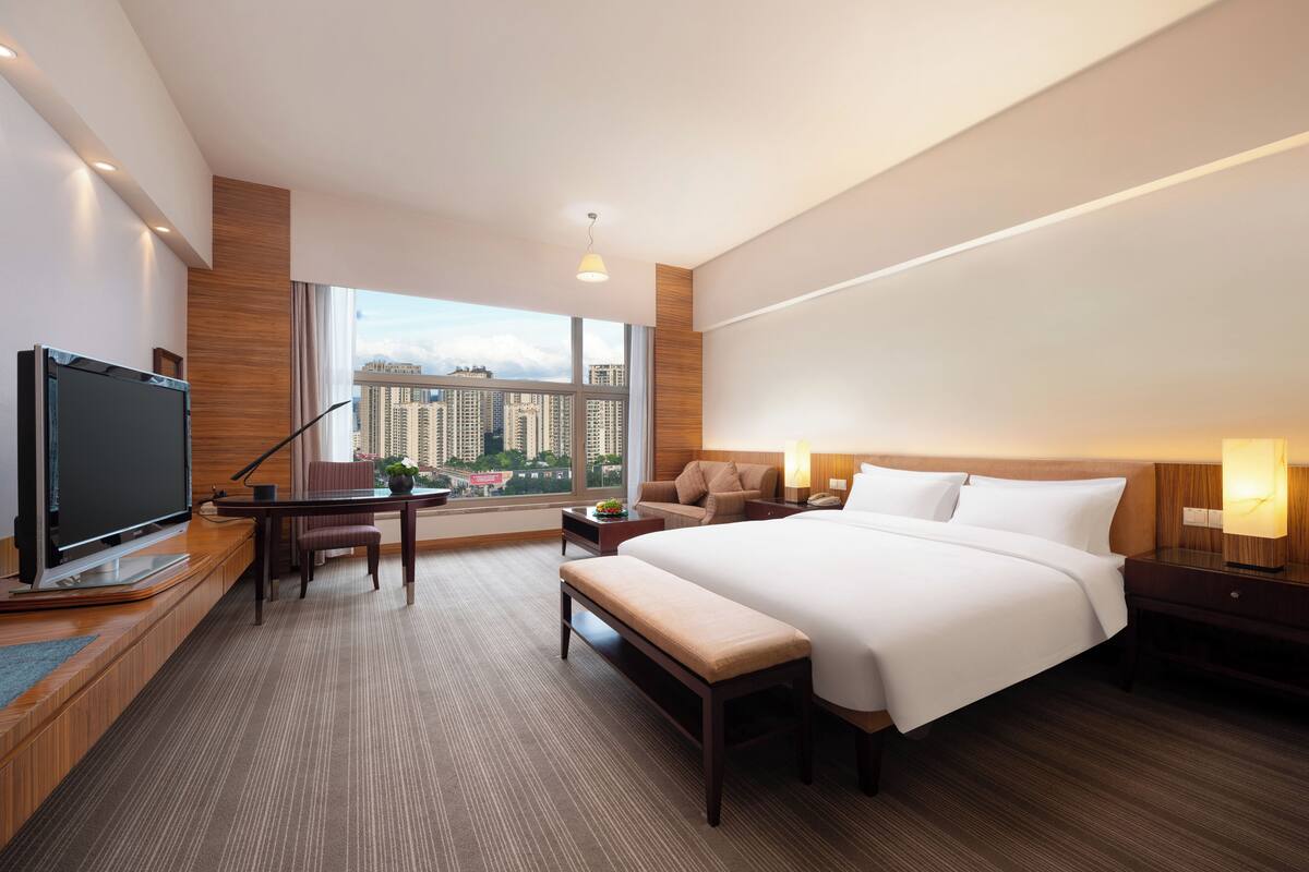 Una habitación de hotel moderna con una cama grande, un escritorio con silla, televisión y vistas de la ciudad a través de amplios ventanales.