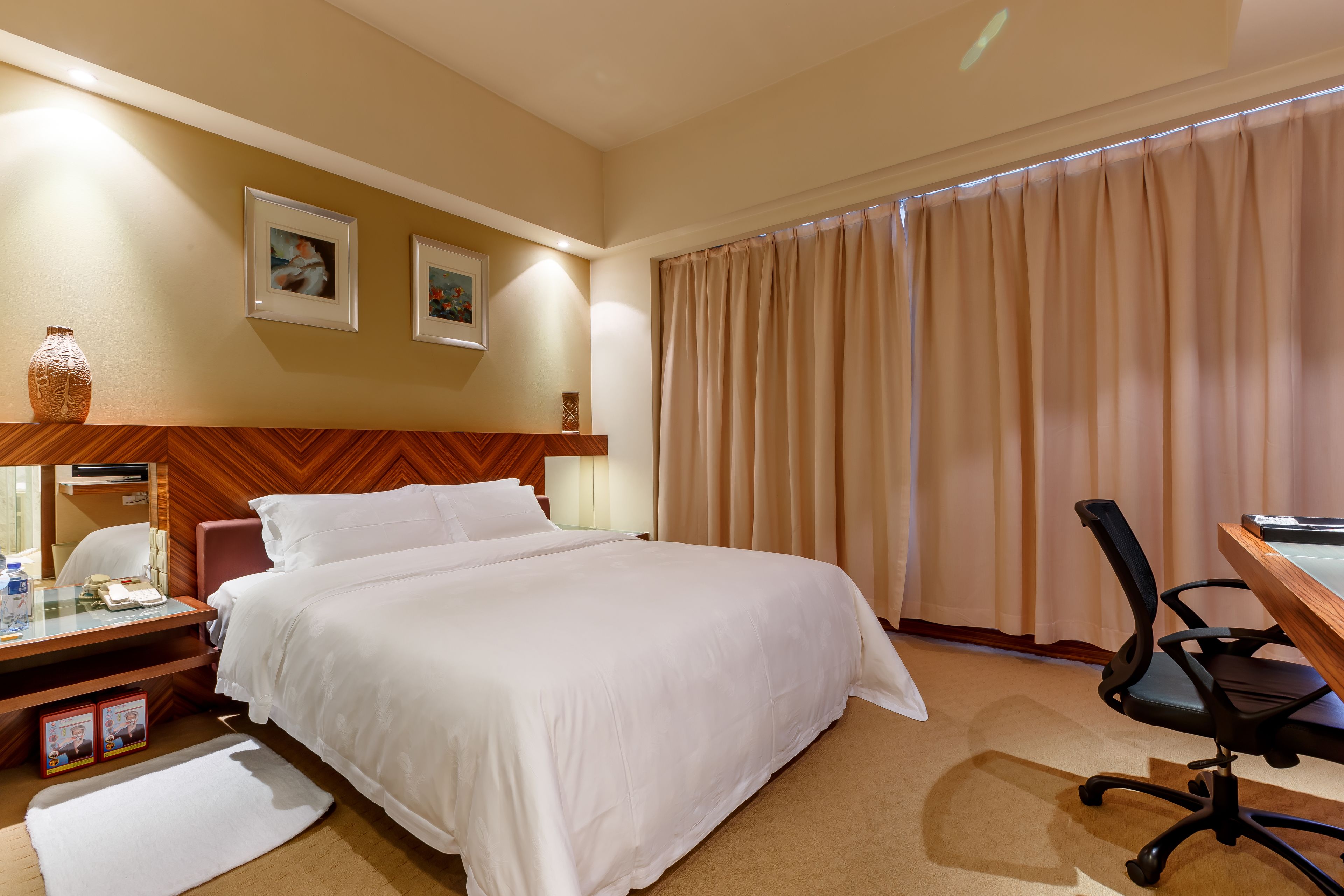 A hotel room with a bed, a desk with a computer, a chair, a lamp, and two framed pictures on the wall.