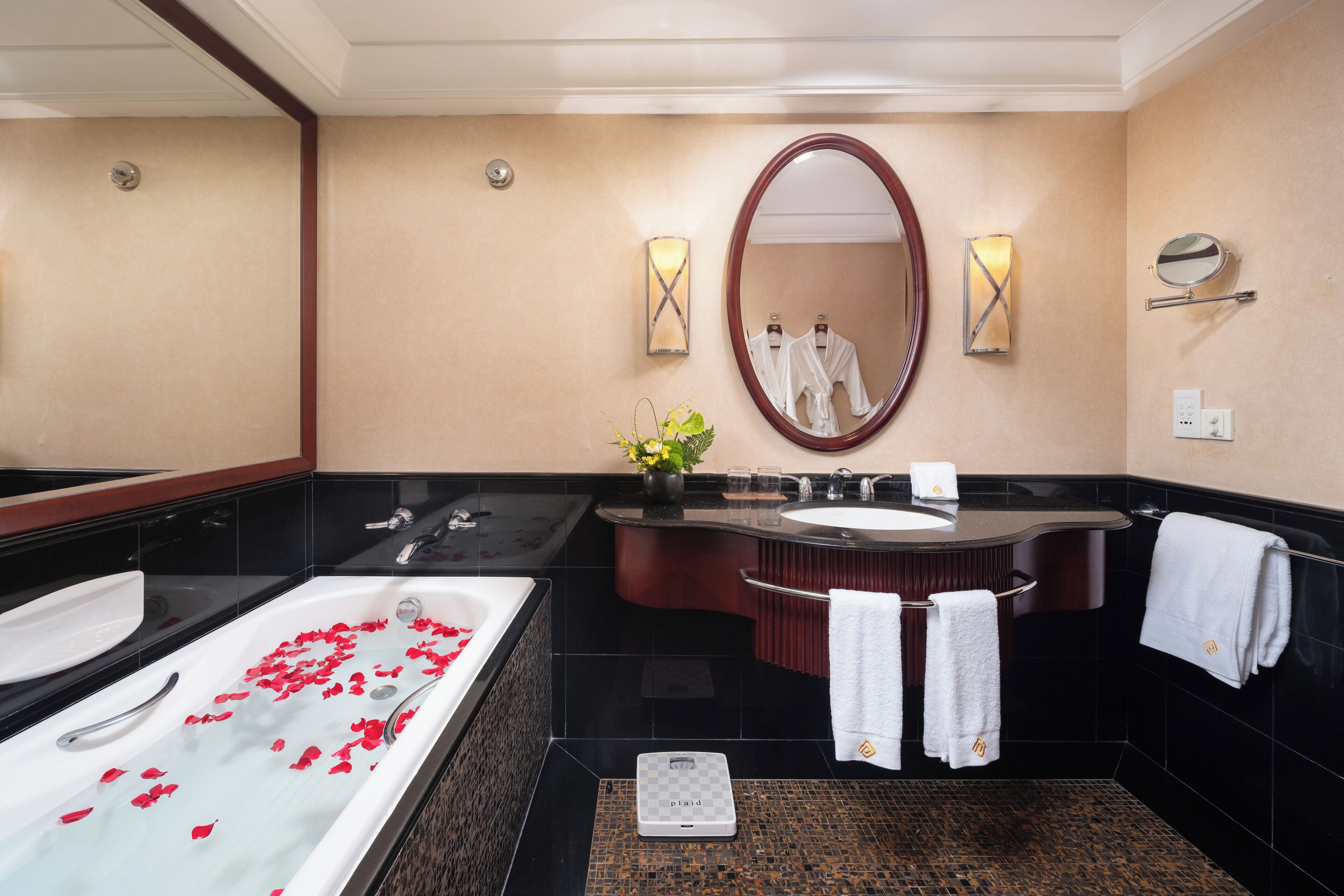 A modern bathroom with two sinks, a large bathtub with rose petals, a scale, and towels hanging on a rack.