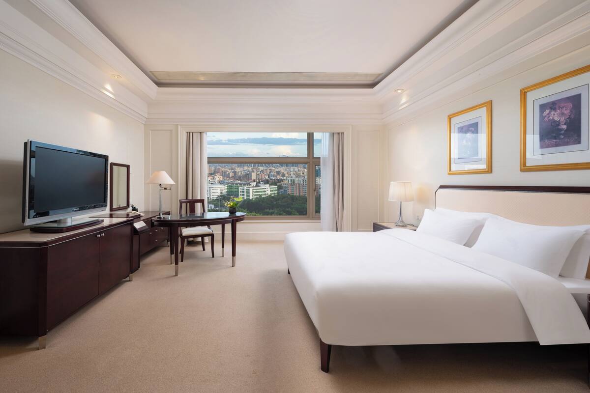 Habitación de hotel con una cama grande, un escritorio con televisor, una silla y vistas a la ciudad a través de un ventanal.
