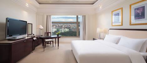 A hotel room with a large bed, a desk with a TV, a chair, and a view of the city through a large window.