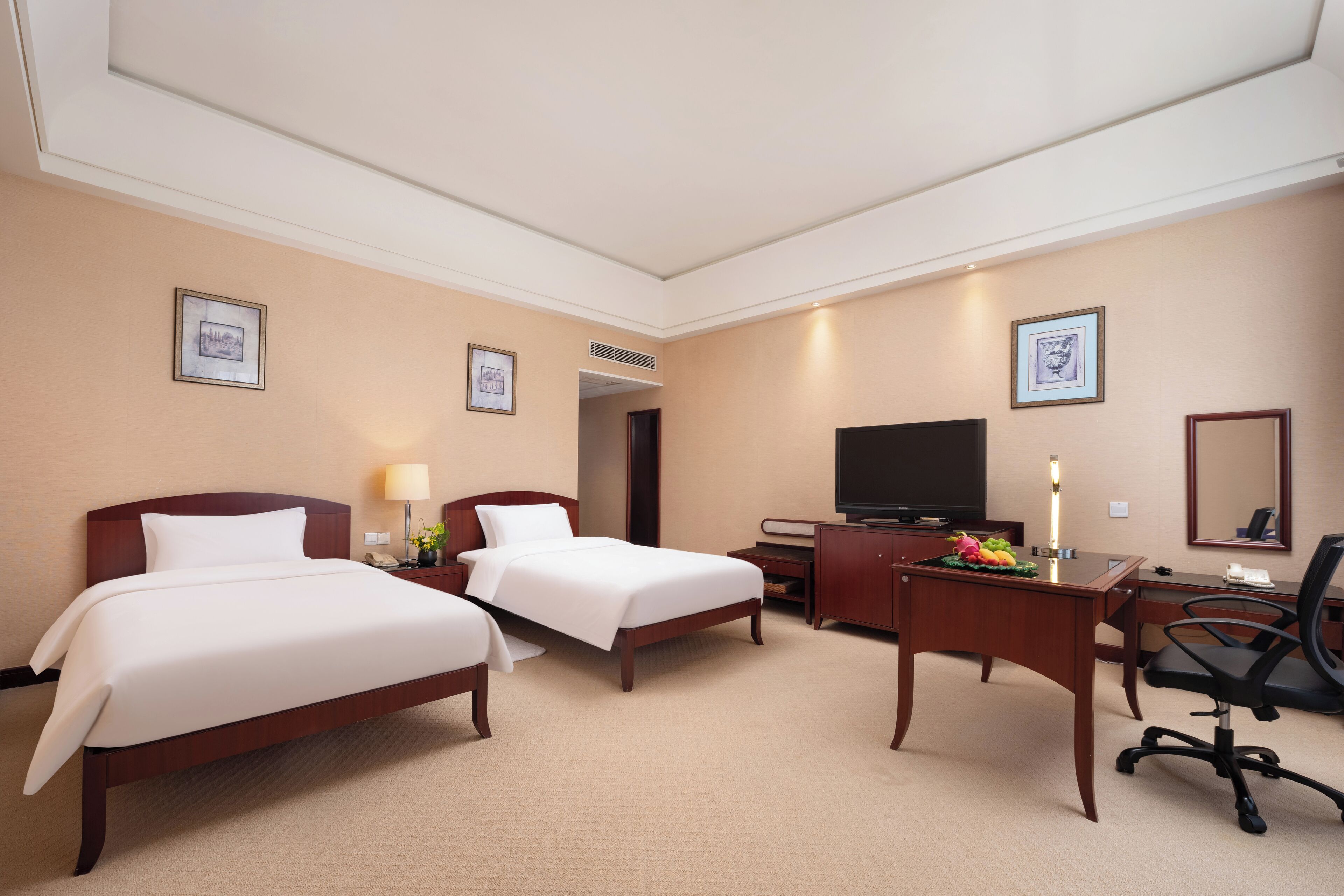 Habitación de hotel con dos camas, un escritorio, una silla, un televisor y cuadros en las paredes.