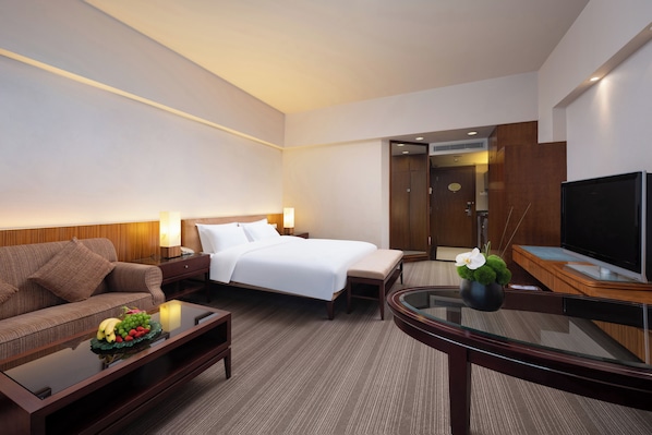 A hotel room with a bed, a sofa, a TV, a coffee table, and a small plant. - Parklane Hotel (Dongguan)