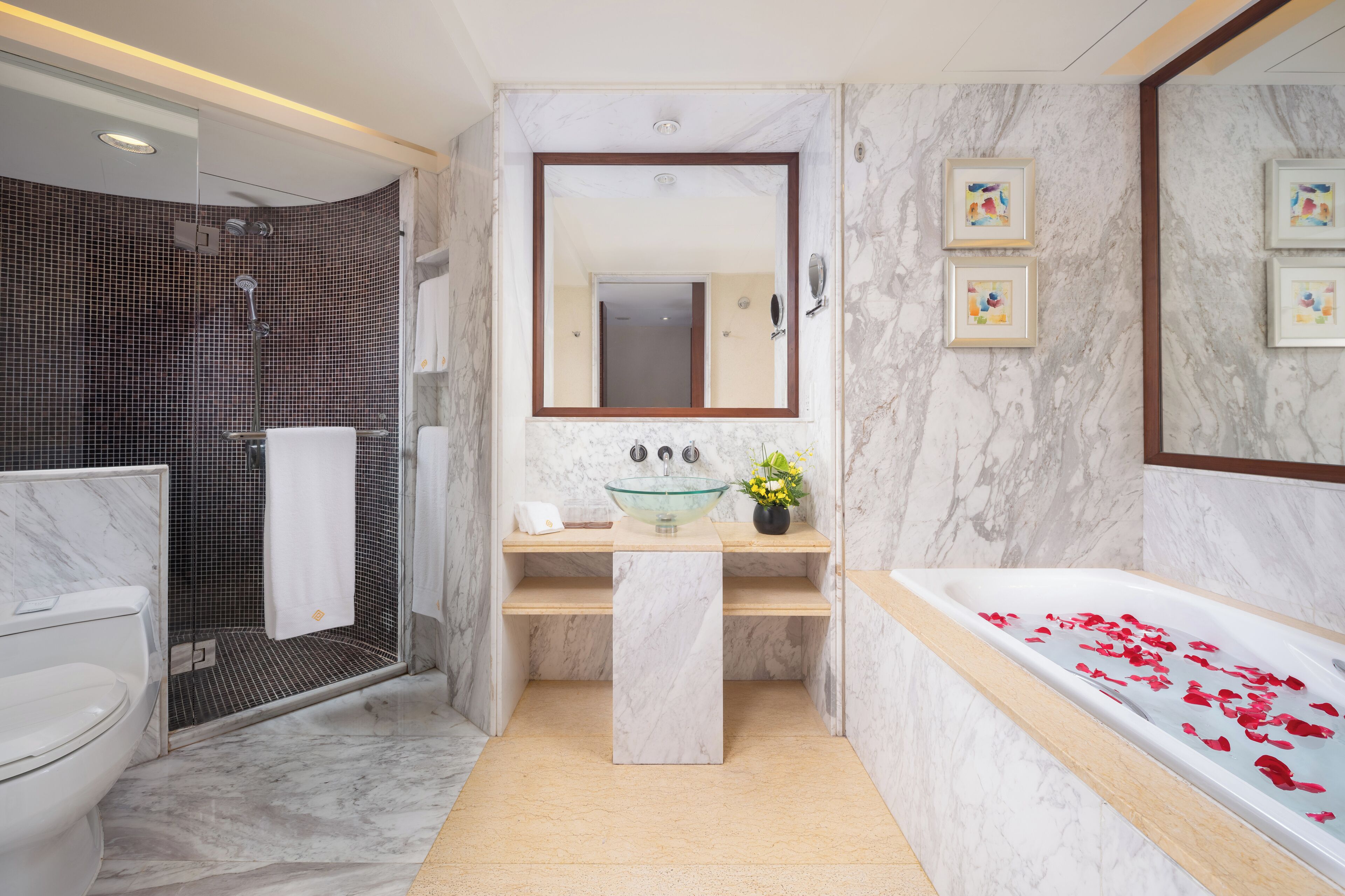 A modern bathroom with a glass sink, a bathtub with rose petals, a shower, and a marble wall.