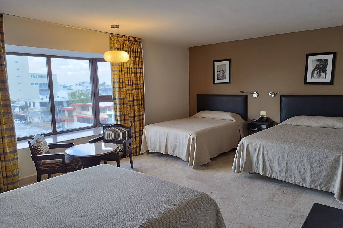 A hotel room with two beds, a small table with a chair, a nightstand with a lamp, and a window with curtains.