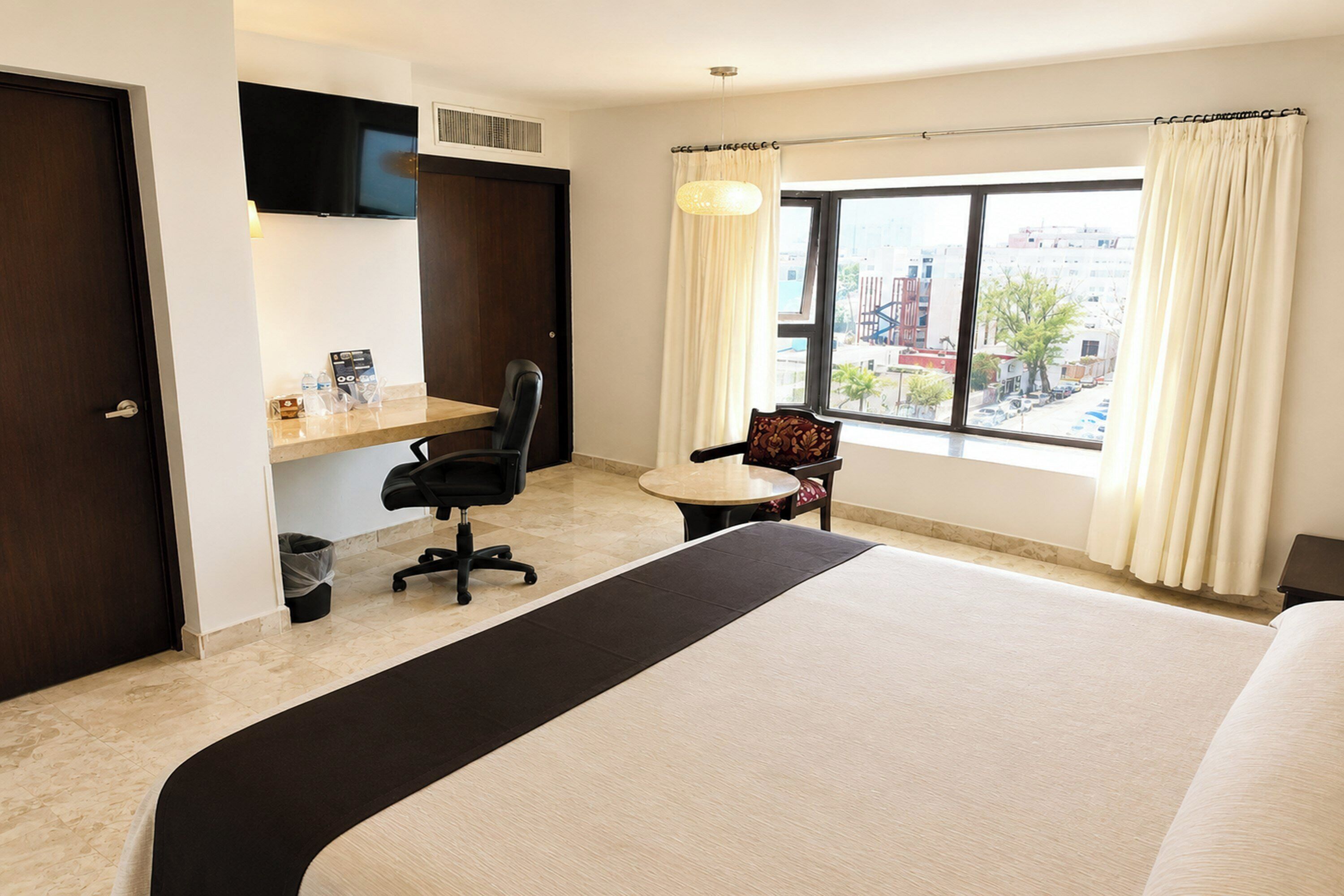 Een hotelkamer met een bed, een bureau met stoel, een raam met gordijnen en uitzicht op de stad.