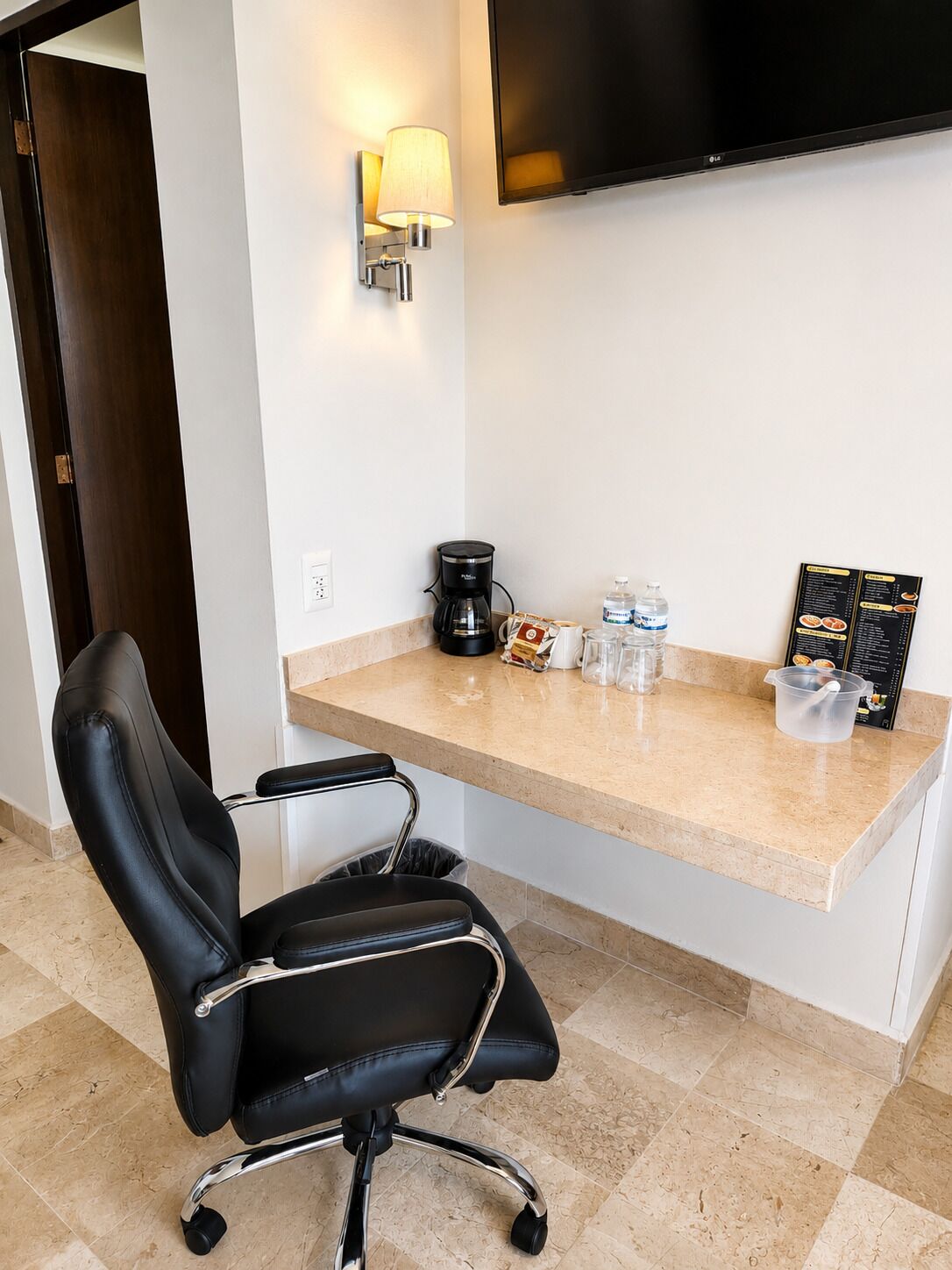 A hotel room with a black leather chair, a coffee maker, a water bottle, a glass pitcher, and a menu board.