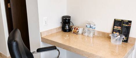 A hotel room with a black leather chair, a coffee maker, a water bottle, a glass pitcher, and a menu board.