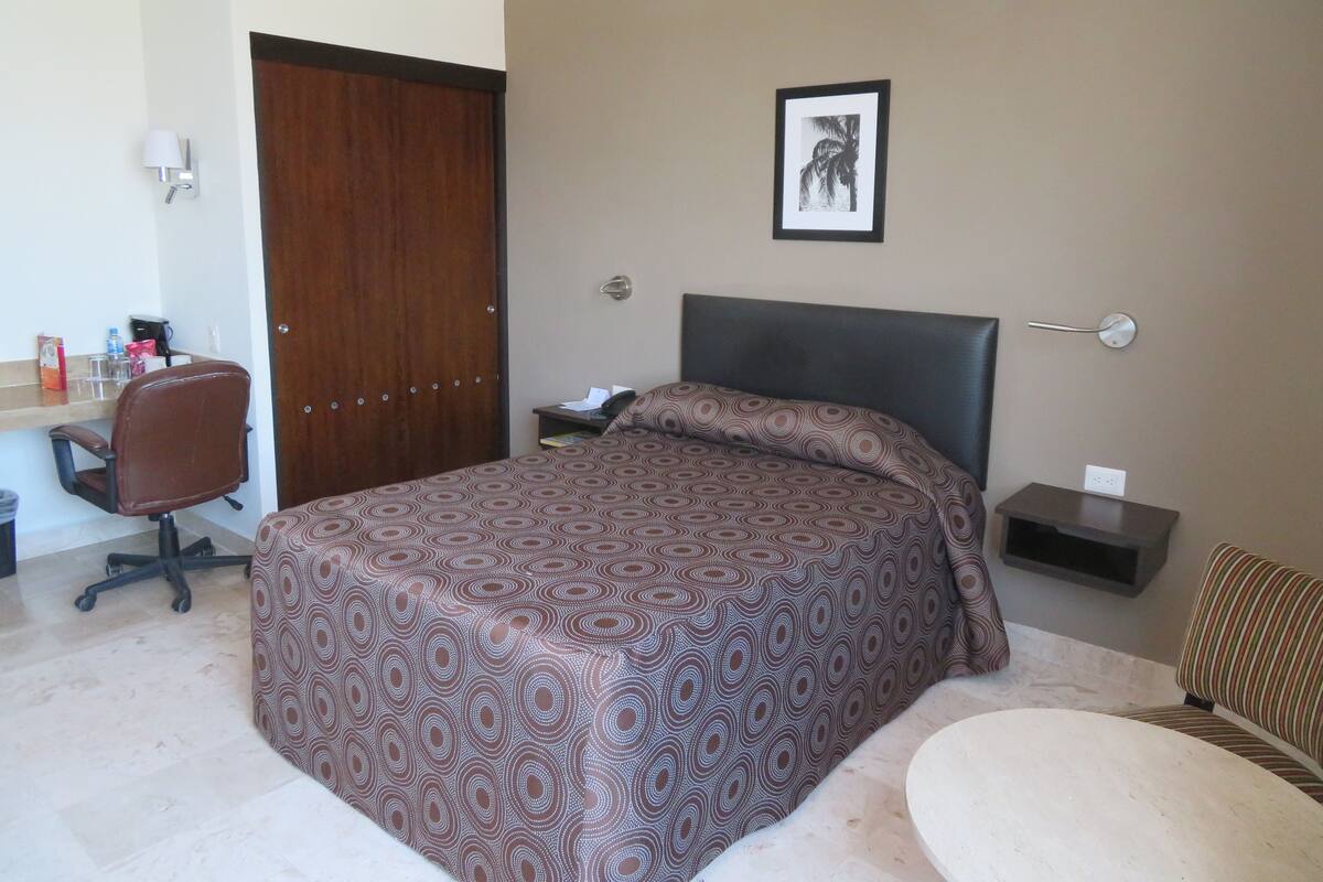 A hotel room with a bed, a desk with a chair, a small bedside table, and a framed picture on the wall.