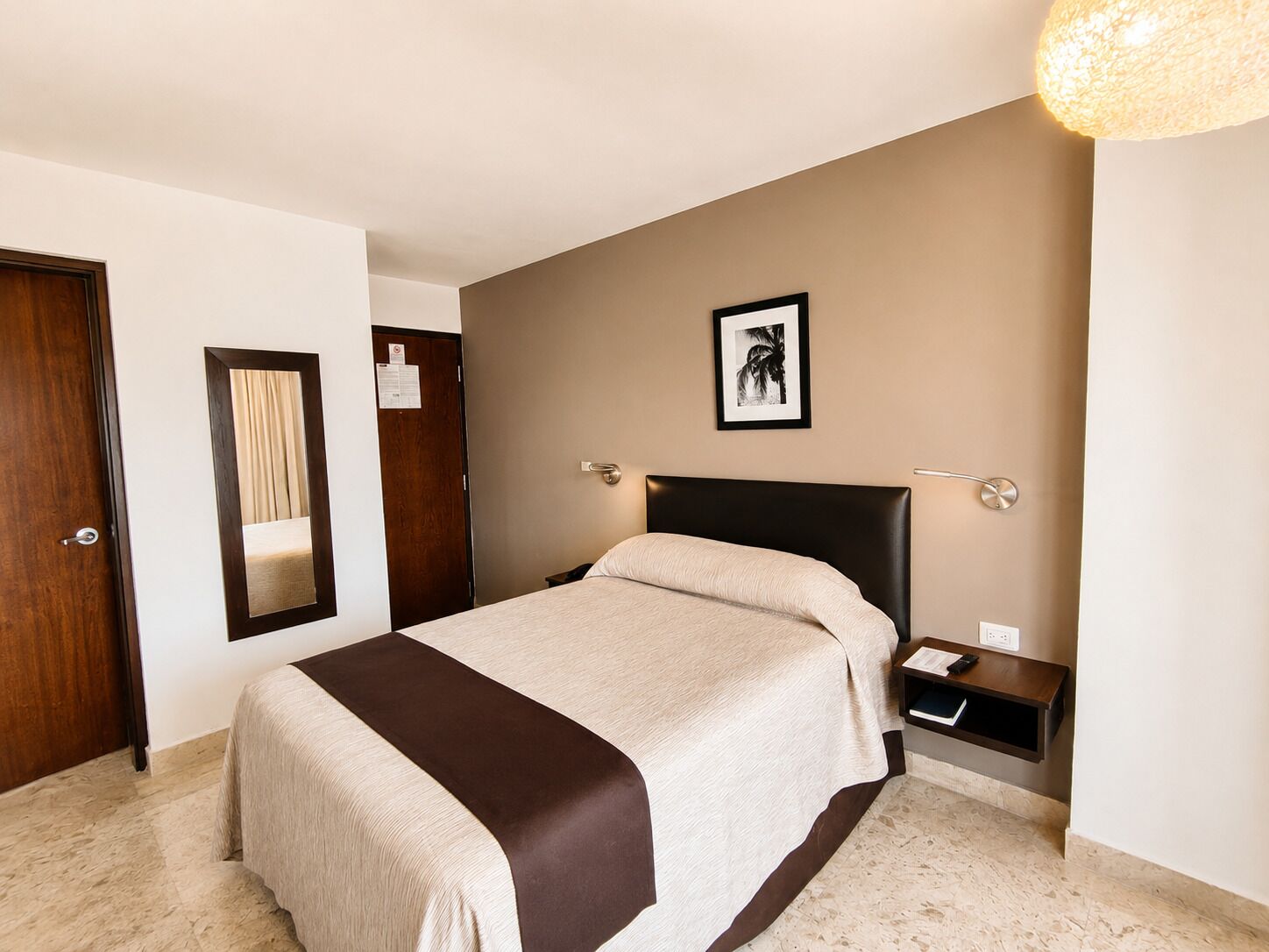 Ein Hotelzimmer mit einem Bett, einem Nachttisch, einer Wandleuchte, einem gerahmten Bild und einer Tür mit Spiegel.
