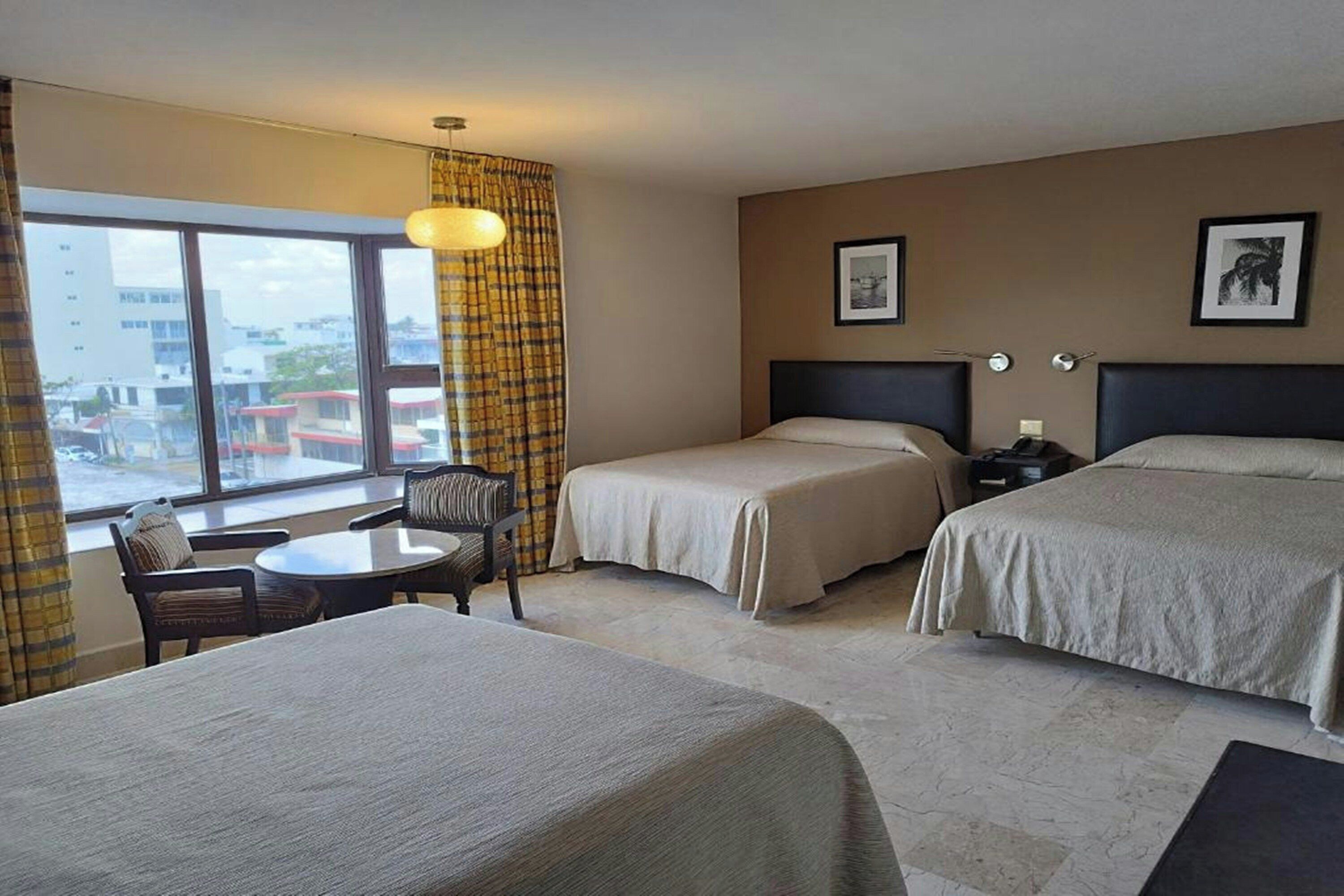 Una habitación de hotel con dos camas, una mesita, una silla y un ventanal con cortinas.