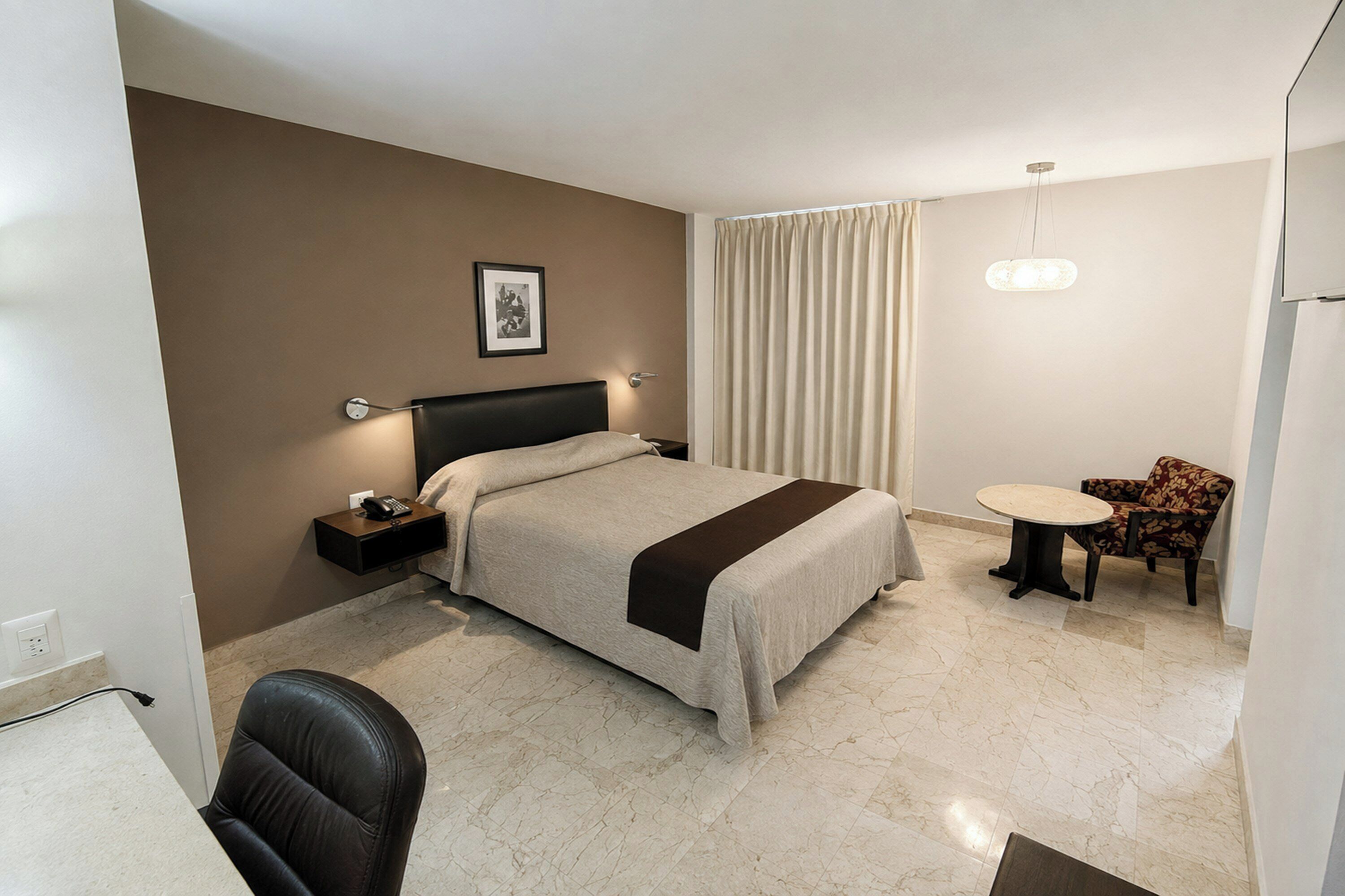 A hotel room with a bed, bedside table, chair, and a small table with a lamp.