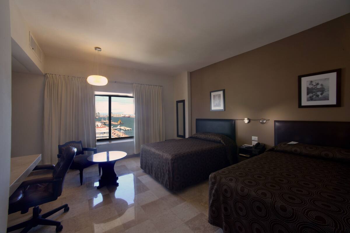A hotel room with a bed, a desk, a chair, a window with curtains, and a framed picture on the wall.