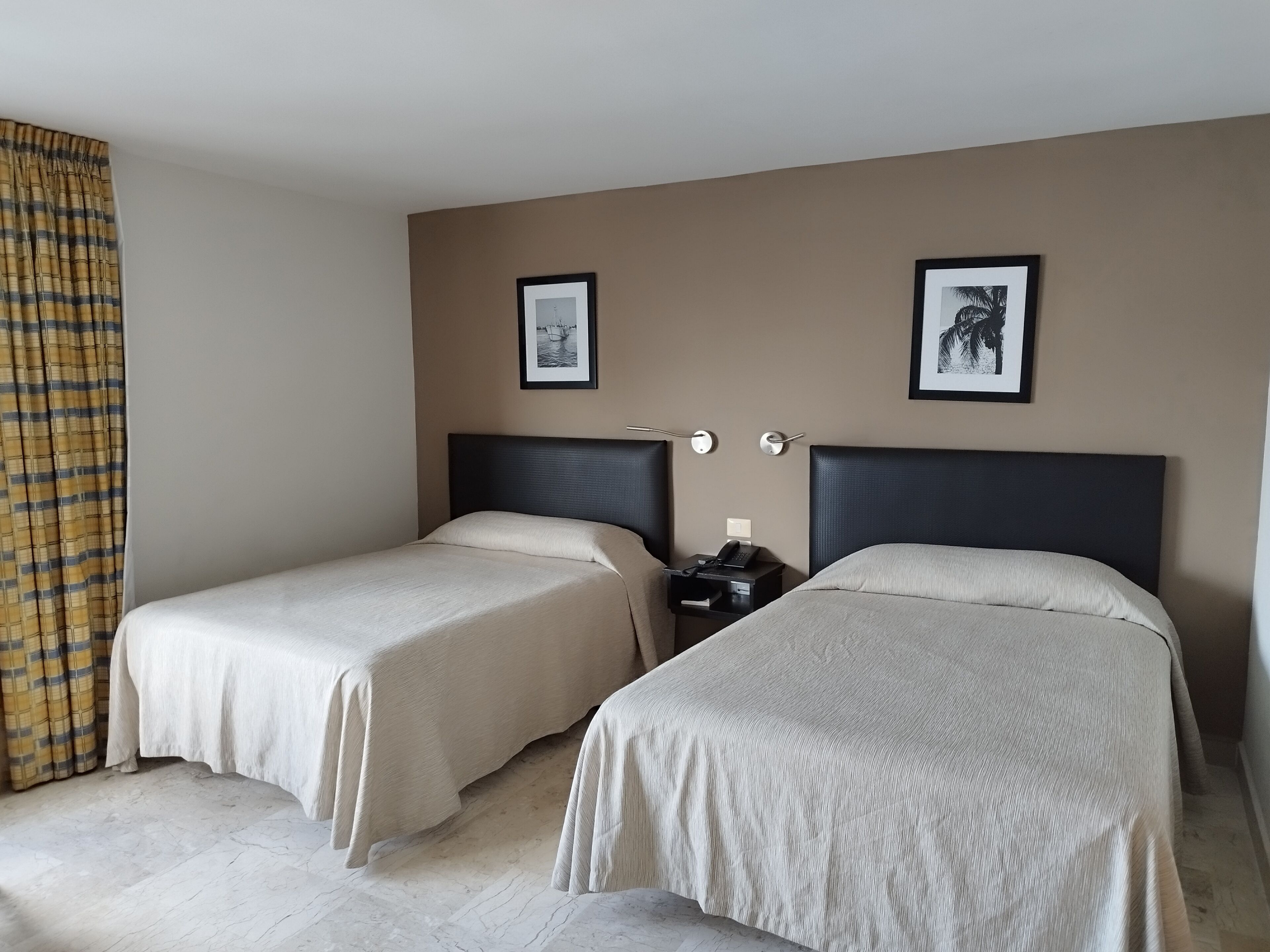 A hotel room with two single beds, a nightstand with a phone, and two framed pictures on the wall.