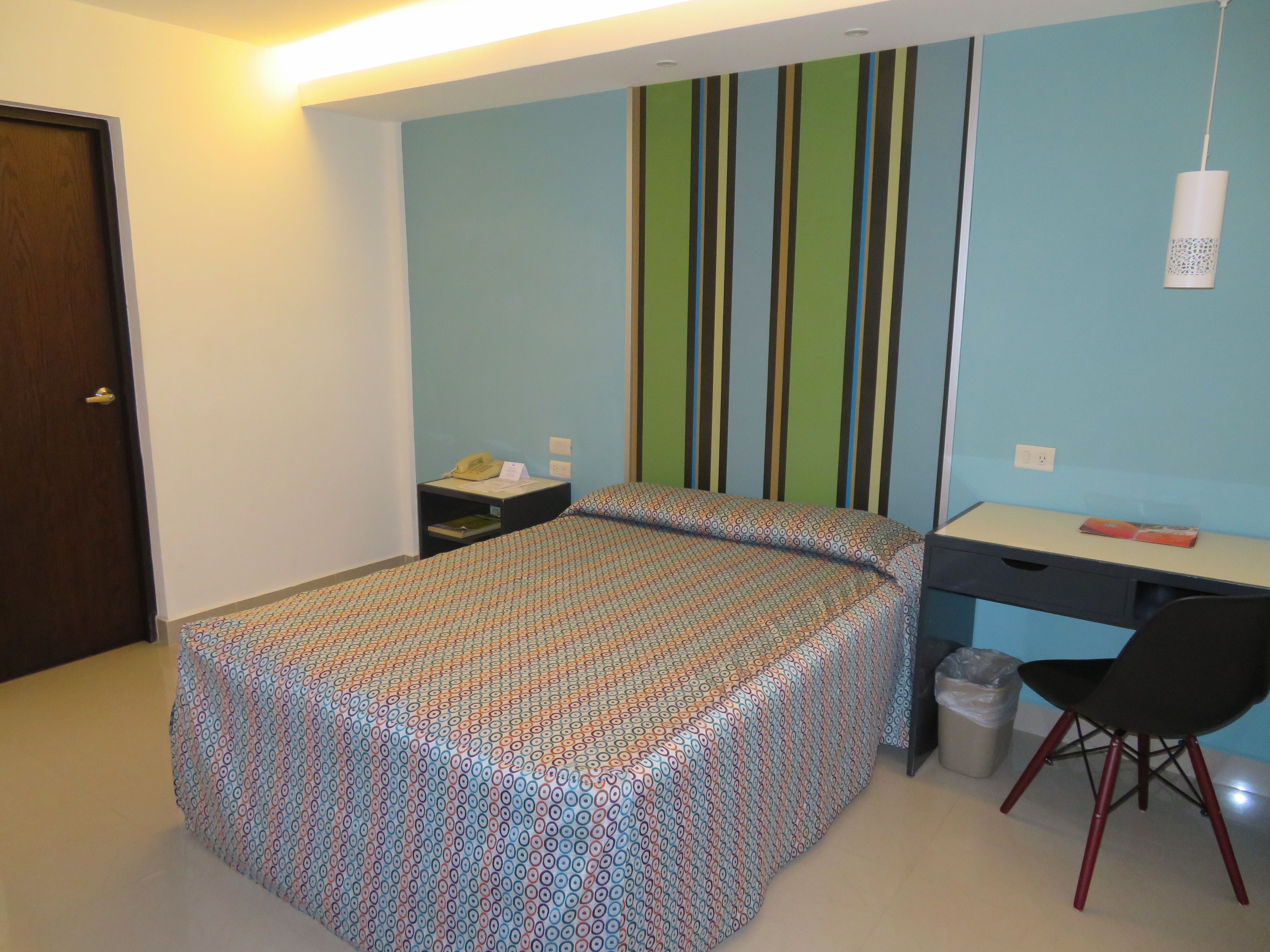 A bedroom with a bed, a desk, a chair, a trash bin, and a wall with vertical stripes.